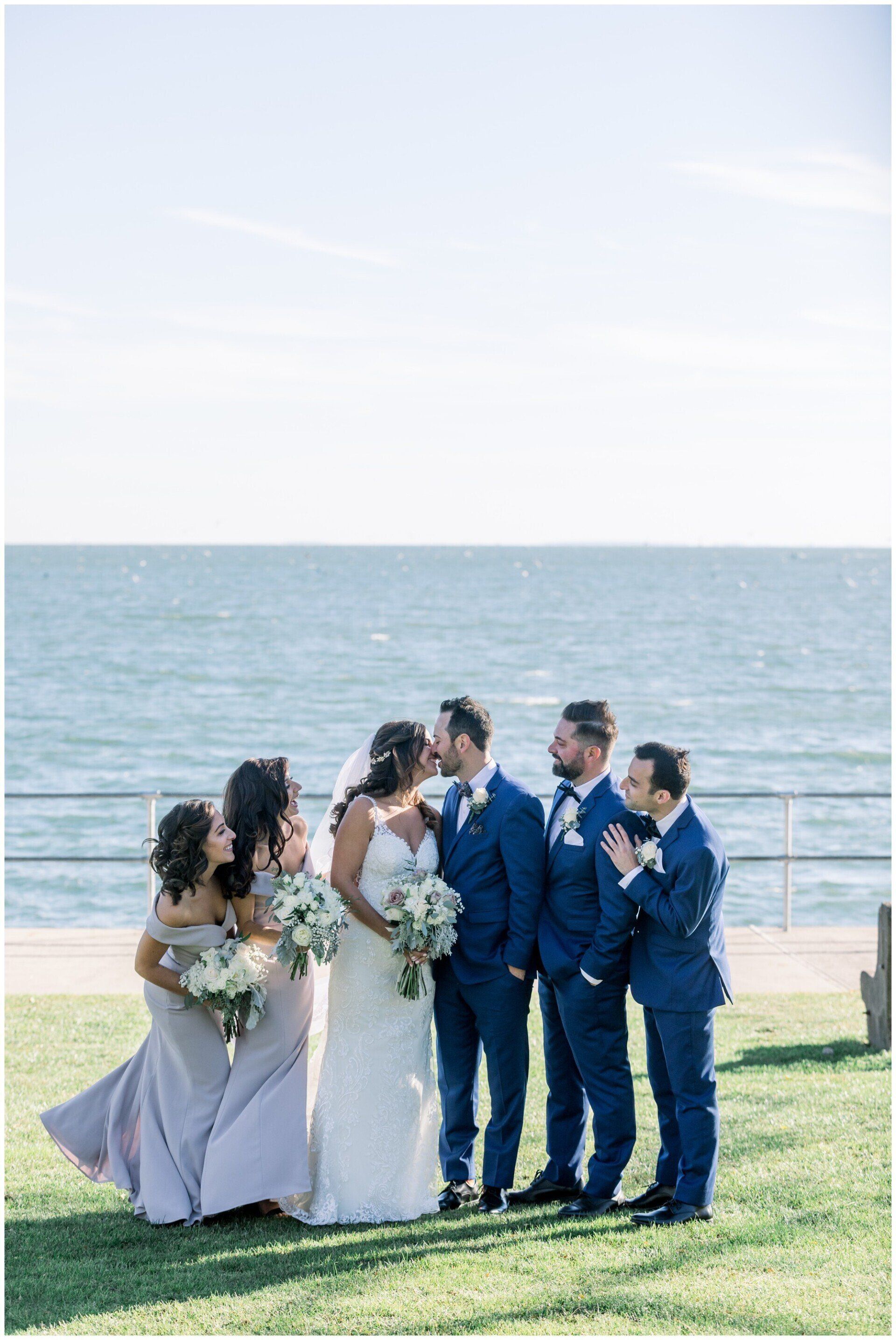 Wedding party posing on a grassy beach with ocean behind them, wearing navy and white attire.