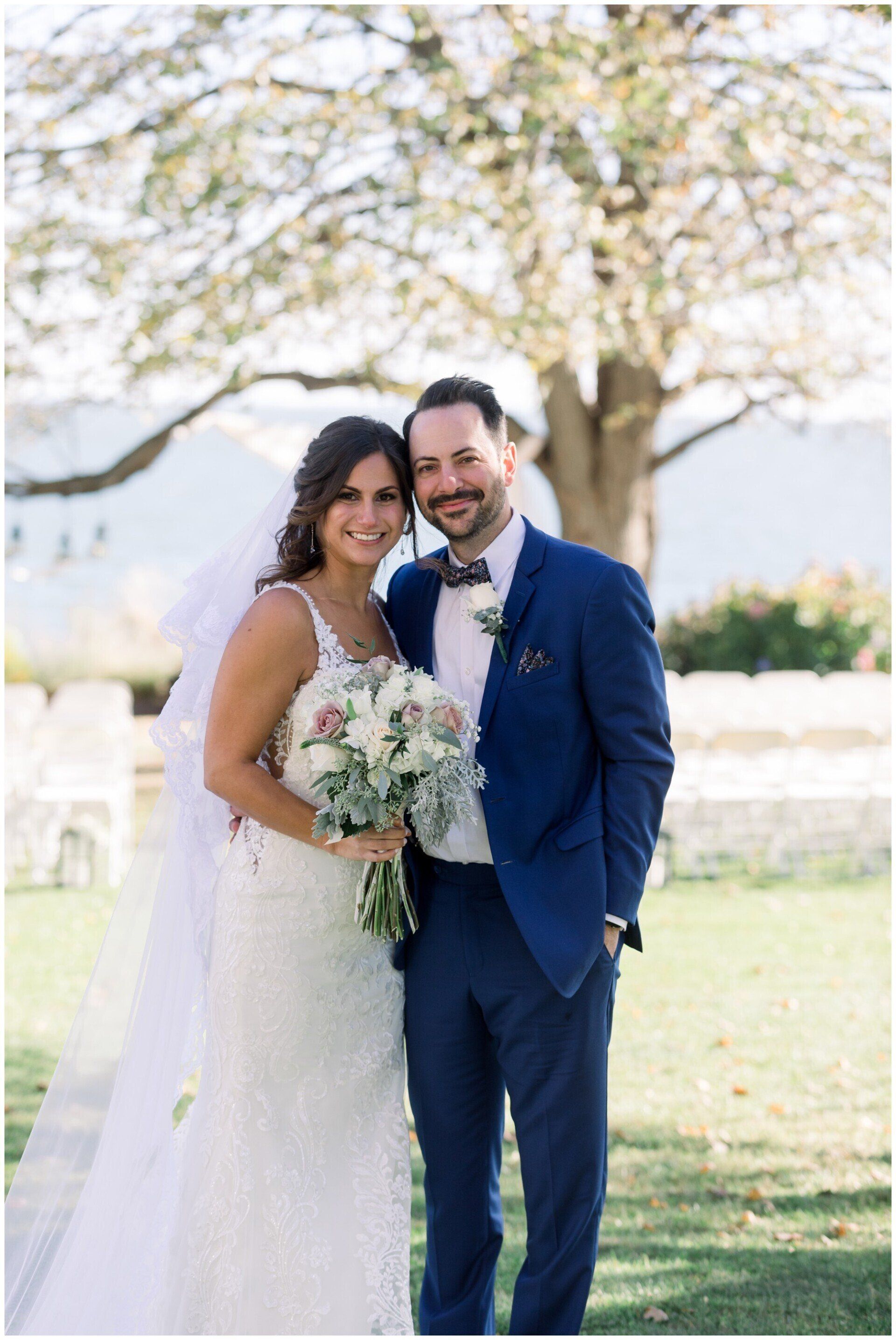 Bride and groom in wedding attire standing outdoors under a blooming tree, smiling and holding a bouquet