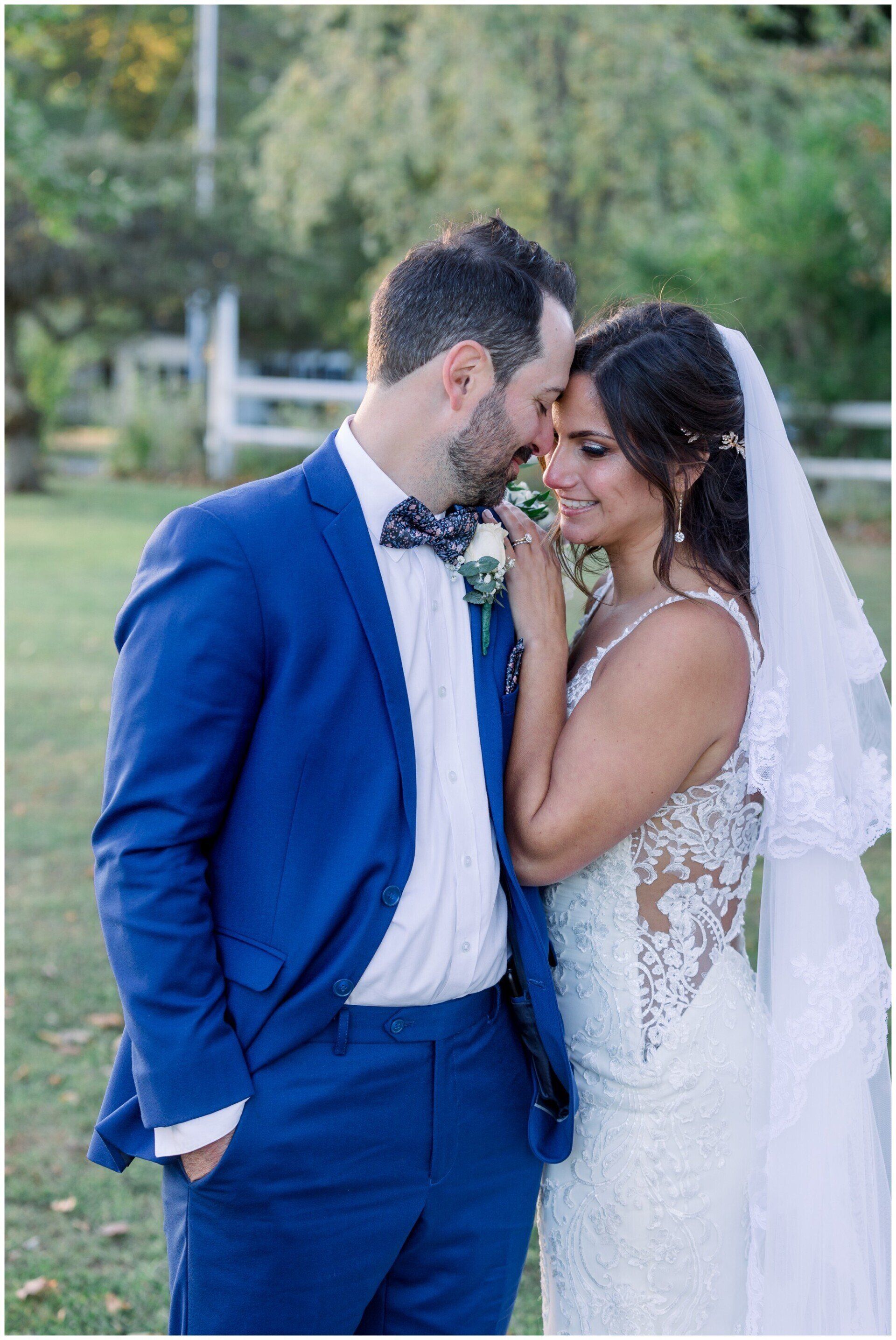 Couple in blue suit and white dress touching foreheads outdoors at a wedding.
