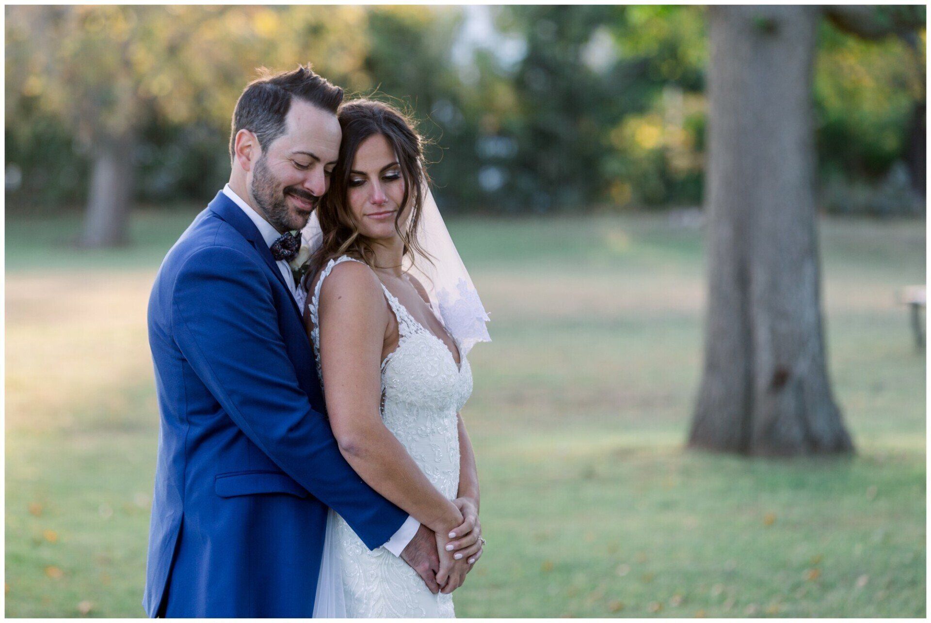 Couple in wedding attire embracing outdoors in a grassy park, with trees in the background