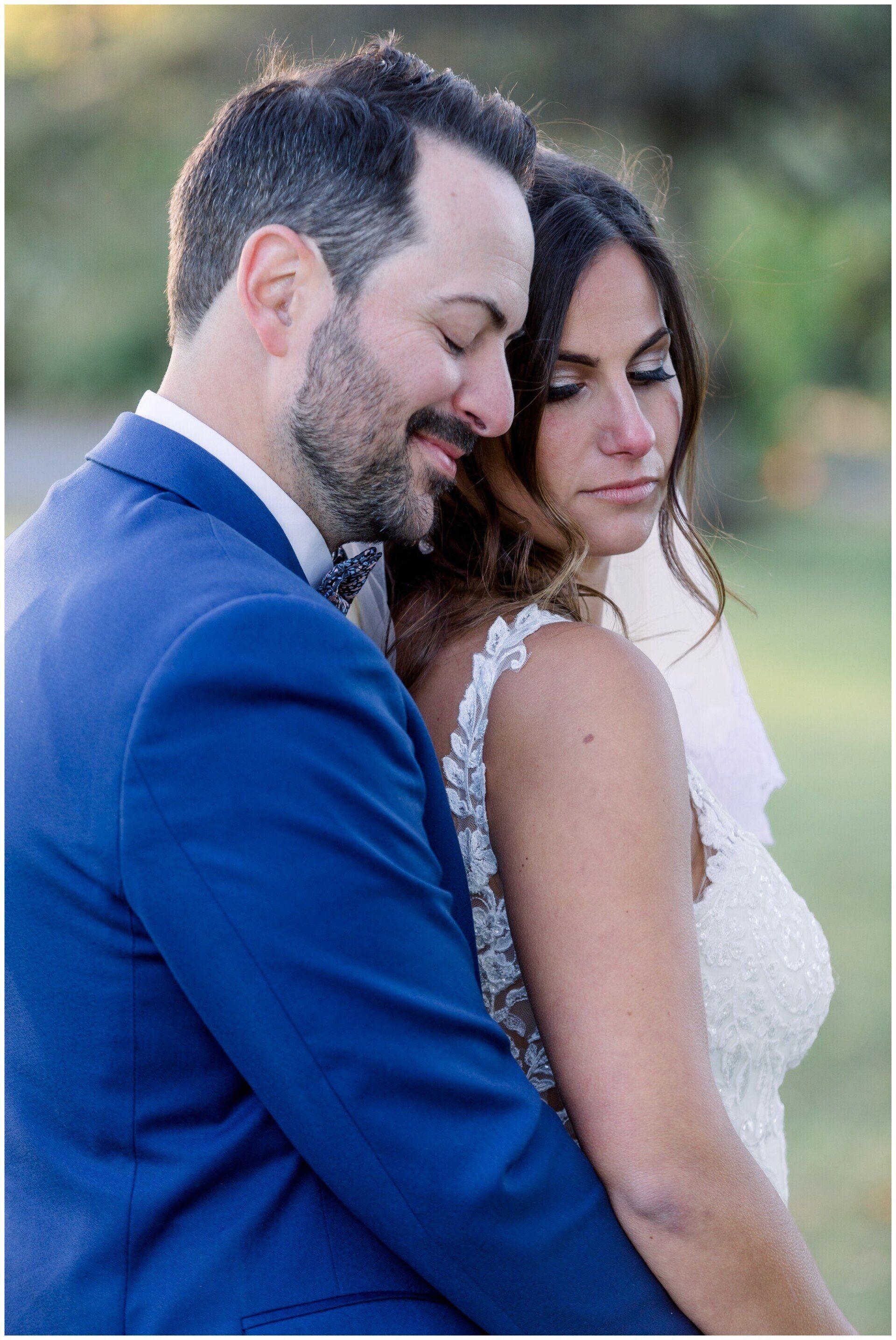 Couple embracing outdoors, man in blue suit resting his head against a woman in a white dress