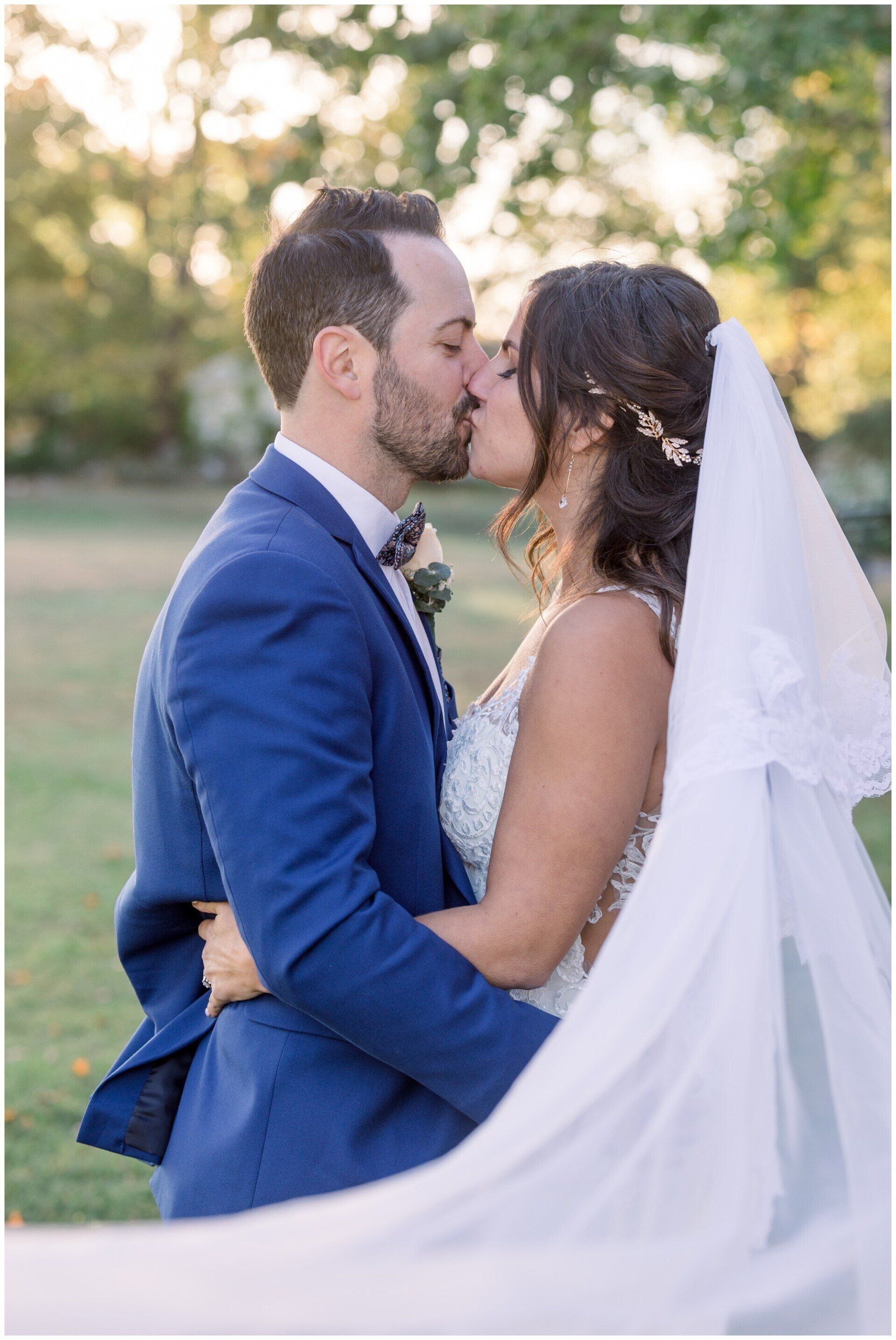Bride and groom in blue suit and white veil kiss outdoors at a wedding