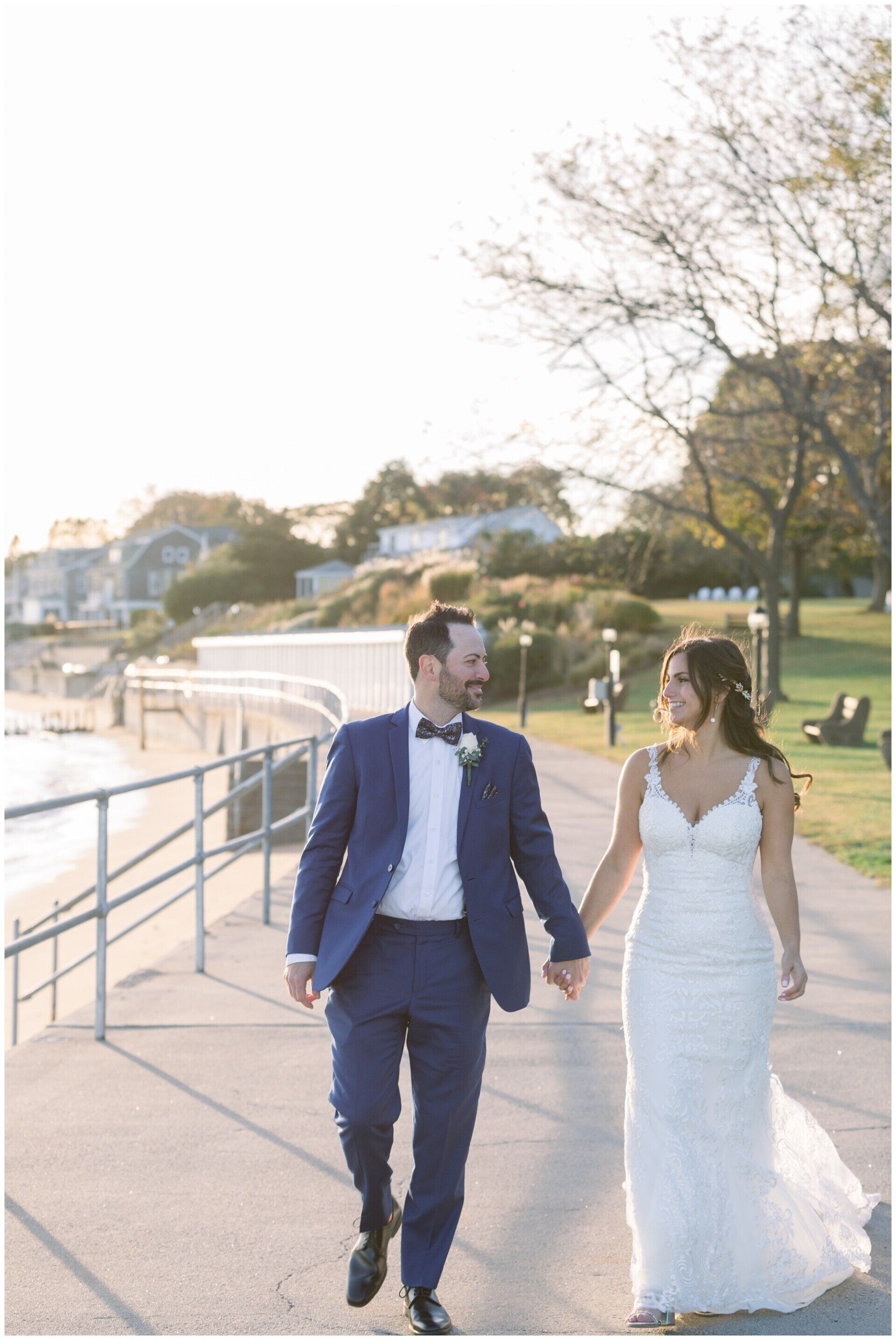 Couple in wedding attire walking hand in hand along a sunlit waterfront path.