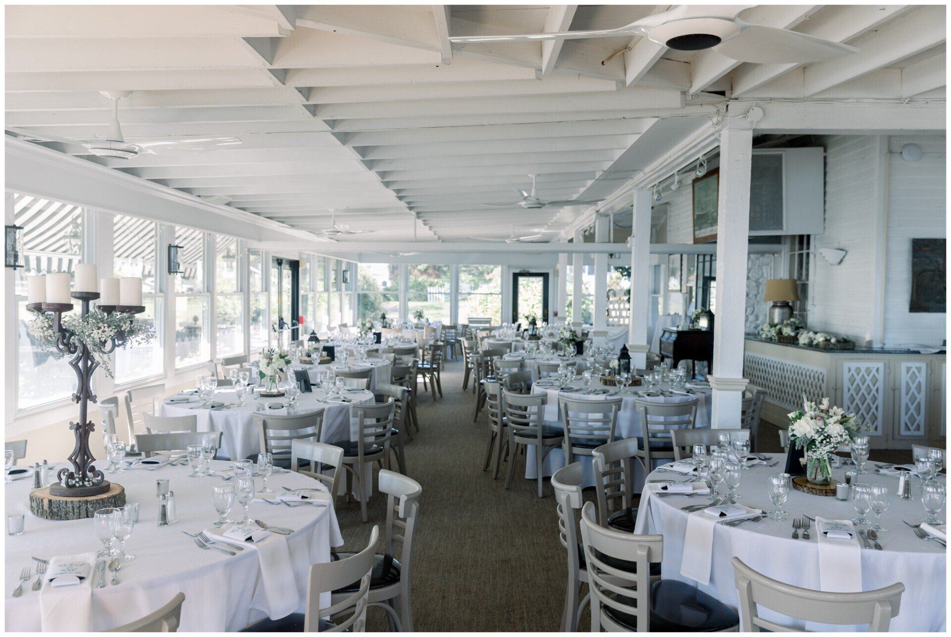 Elegant white-clothed dining room with round tables, chandeliers, and large windows overlooking the water