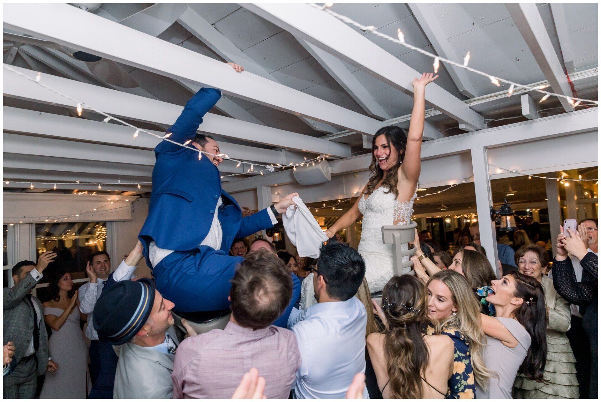 Wedding guests lifting people in the air at a lively indoor reception under white beams