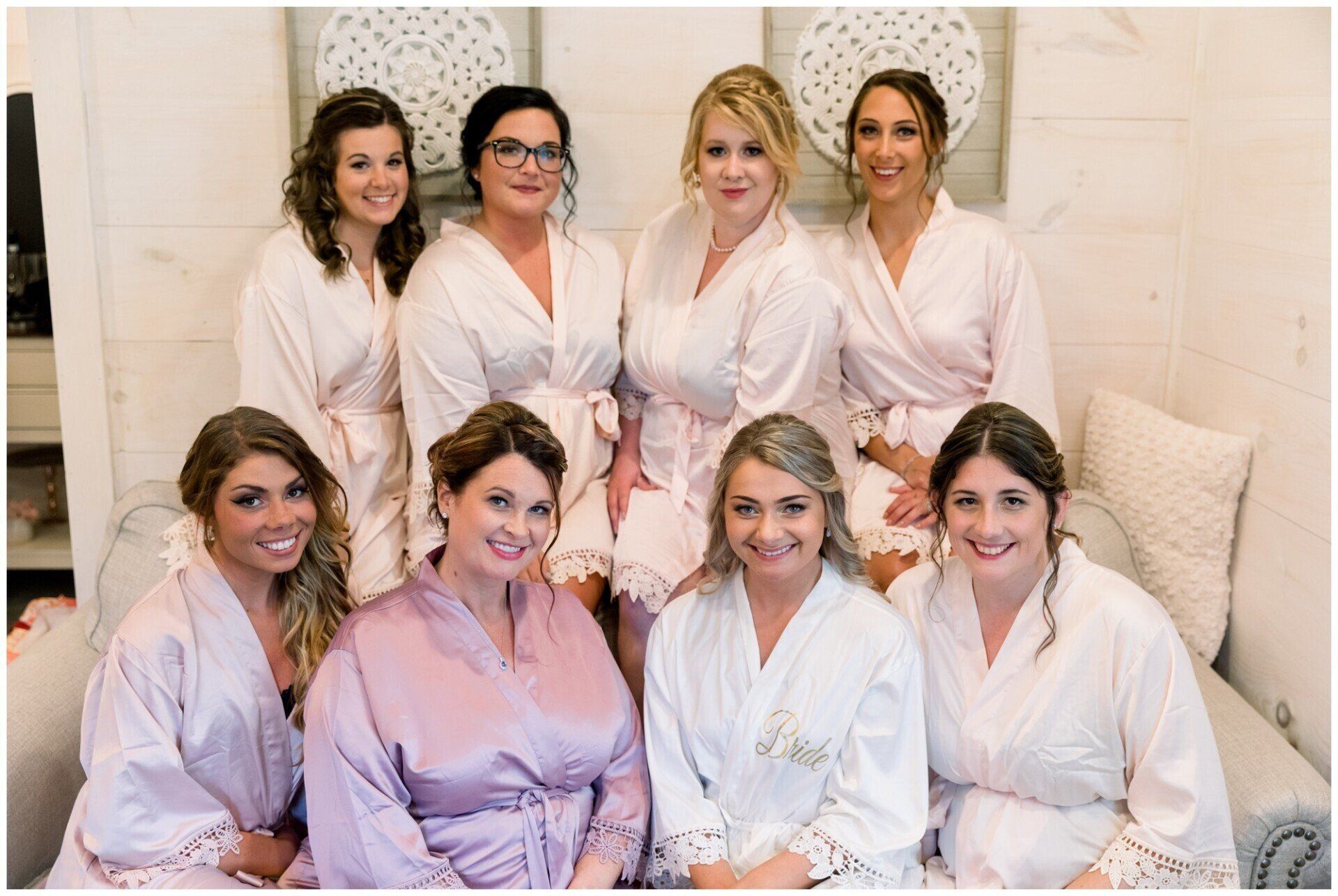 Seven women in white robes smiling together on a sofa in a bright room
