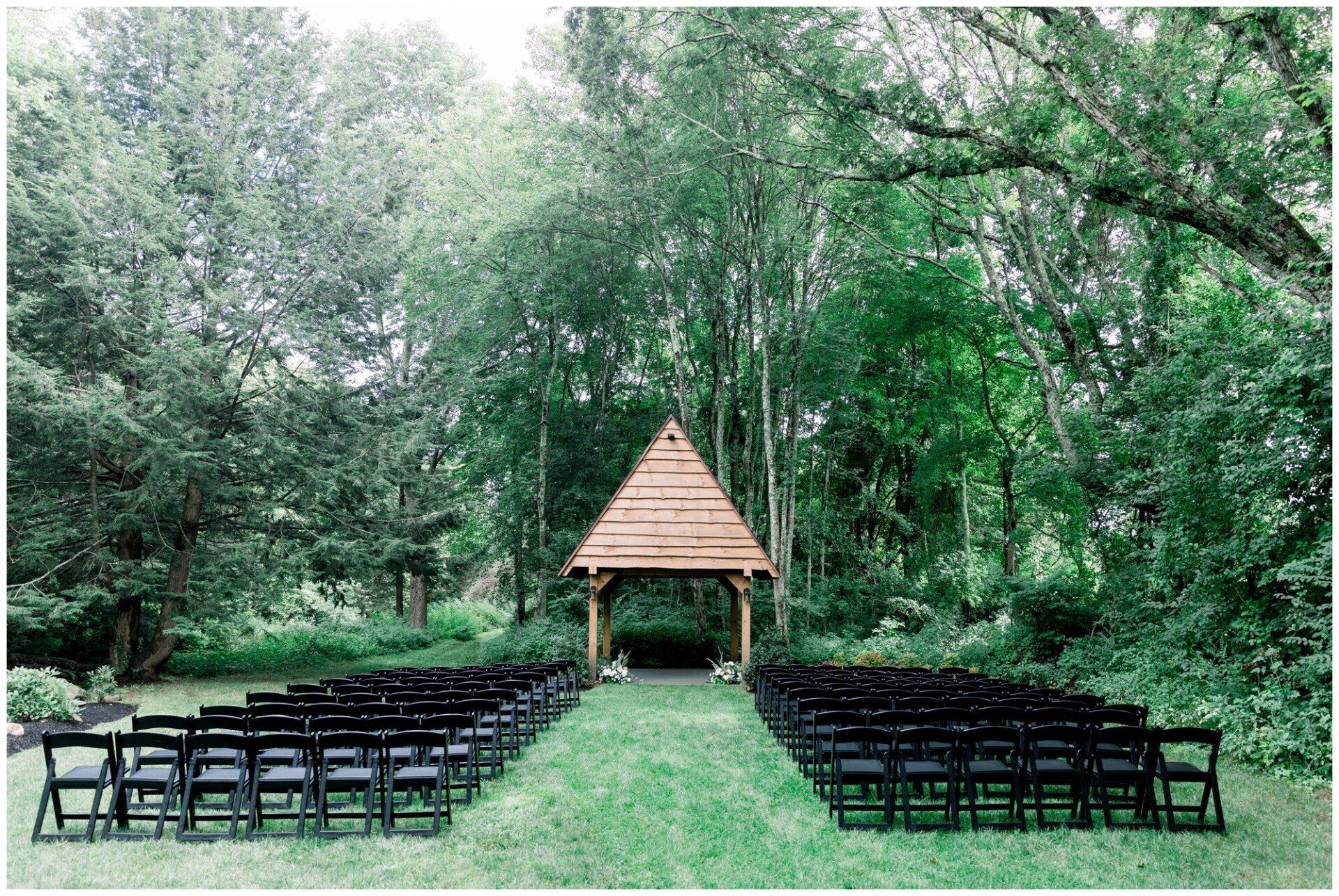 Outdoor wedding ceremony setup with rows of black chairs facing a wooden arch in a lush green forest.