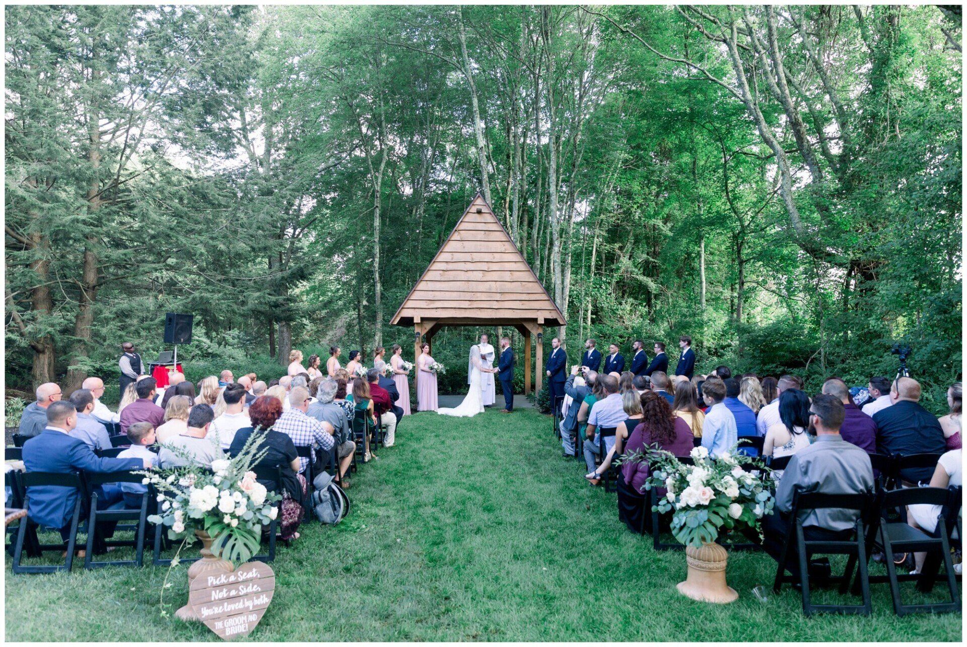 Outdoor wedding ceremony in a wooded grove, with guests seated on white chairs around a small pavilion altar.