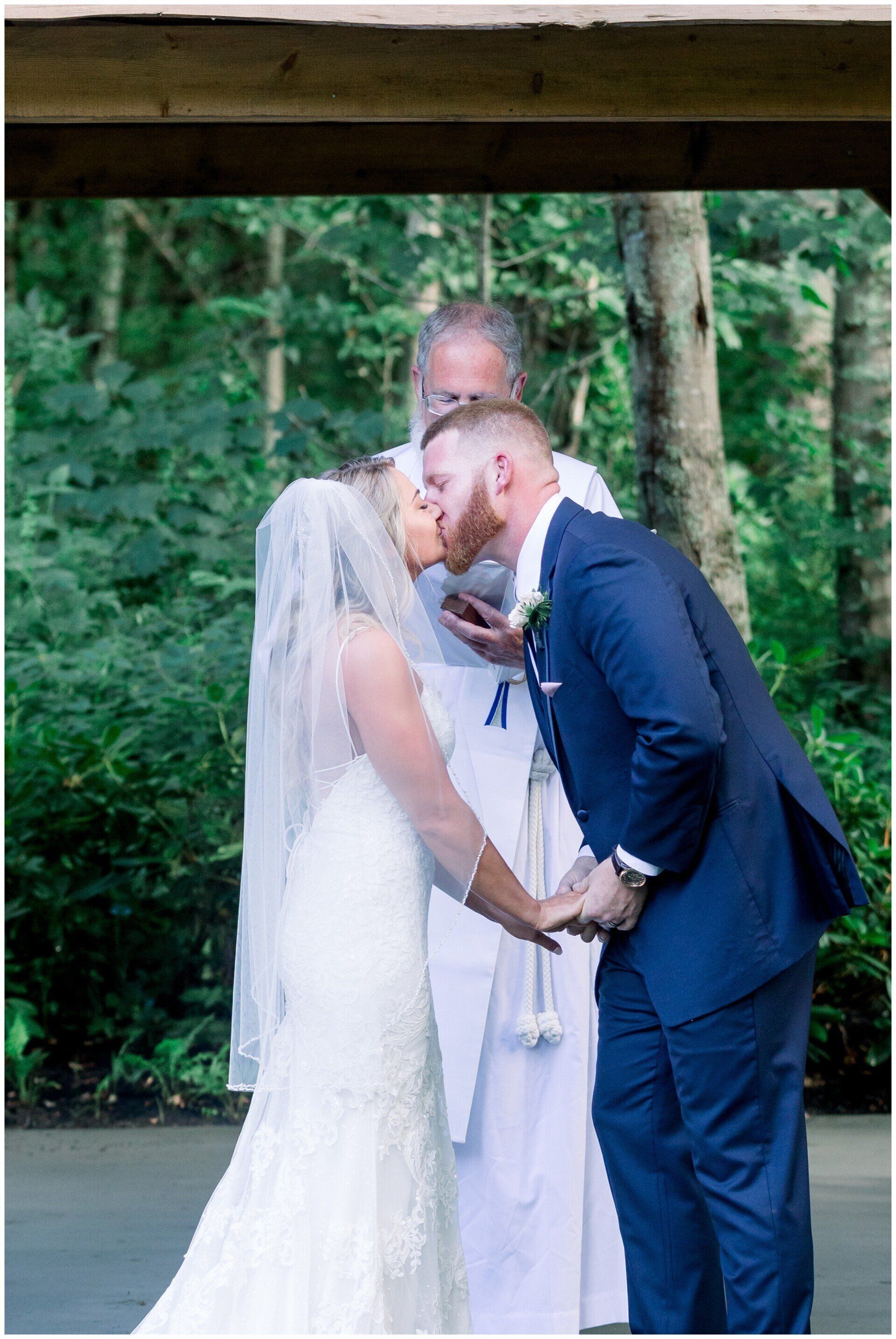 Bride and groom kissing during a wedding ceremony outdoors with trees in the background