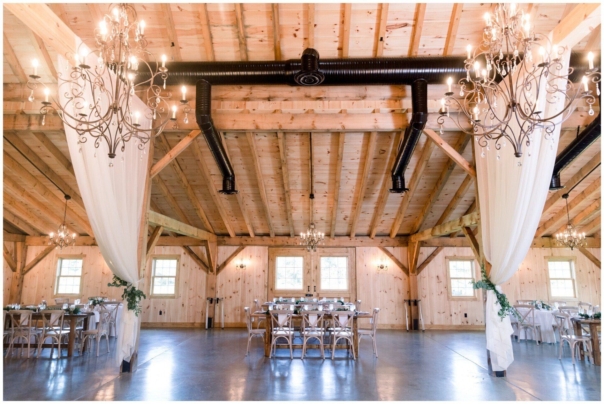 Rustic barn-style event hall with chandeliers, draped white fabric, wooden beams, and rows of chairs