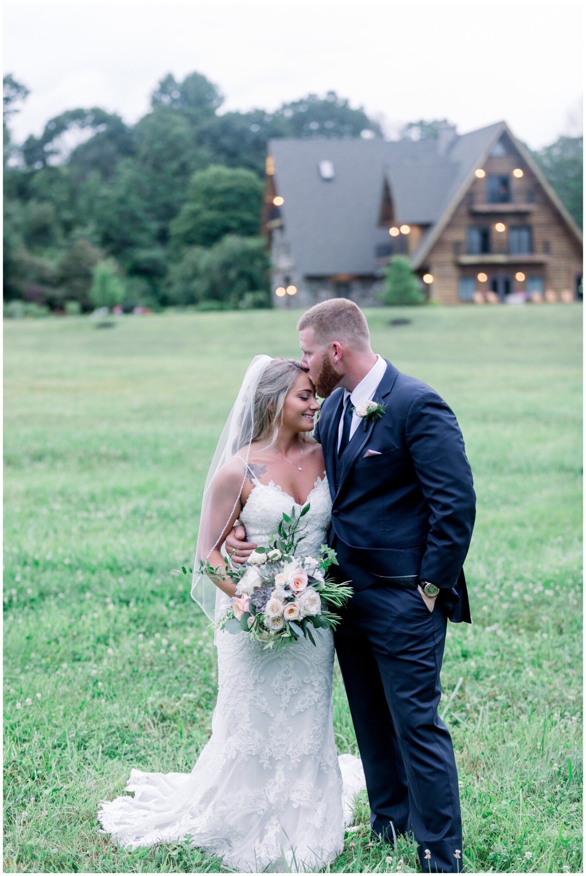 Bride and groom kissing in a grassy field with a rustic house in the background.
