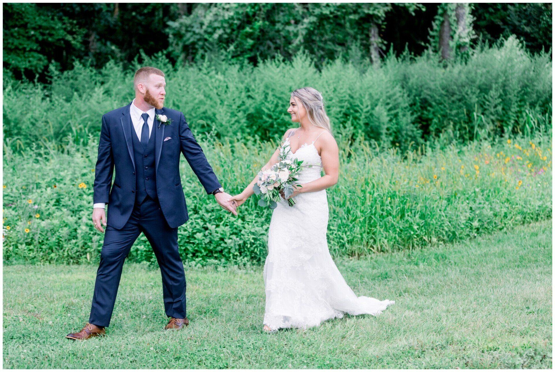 Bride and groom holding hands in a grassy field, with a green wooded backdrop.