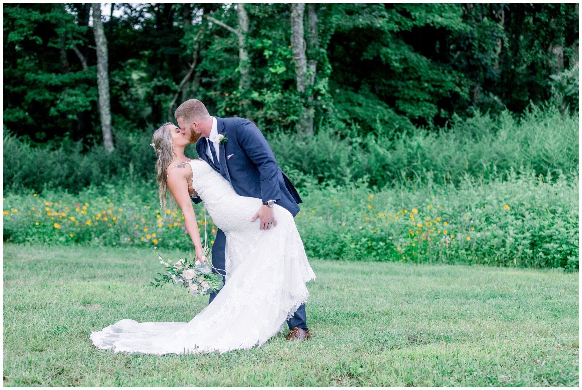 Bride and groom kissing on a grassy lawn, bride in white gown and groom in dark suit, with trees behind them
