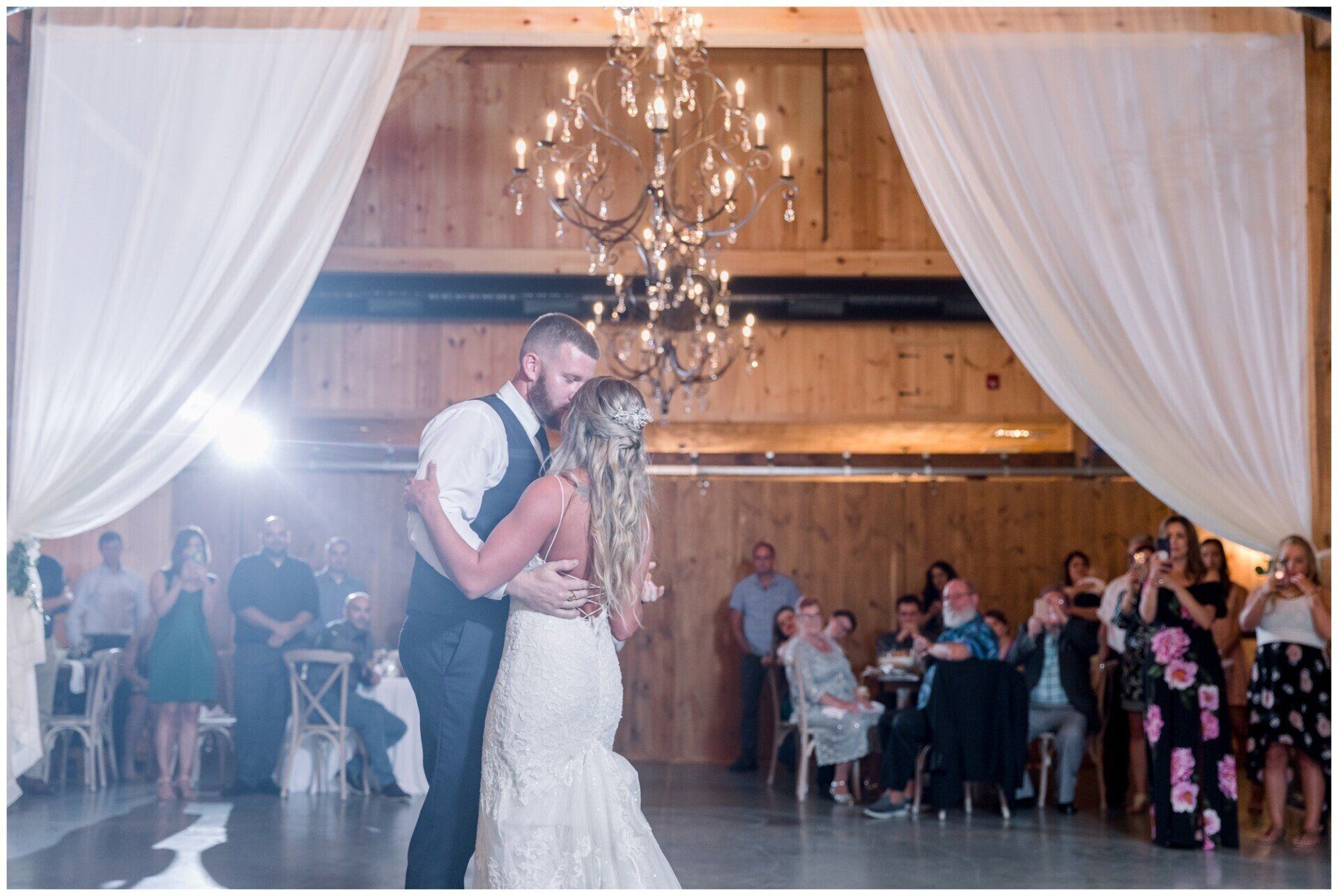 Bride and groom kiss under a chandelier in a rustic wedding reception hall with seated guests.