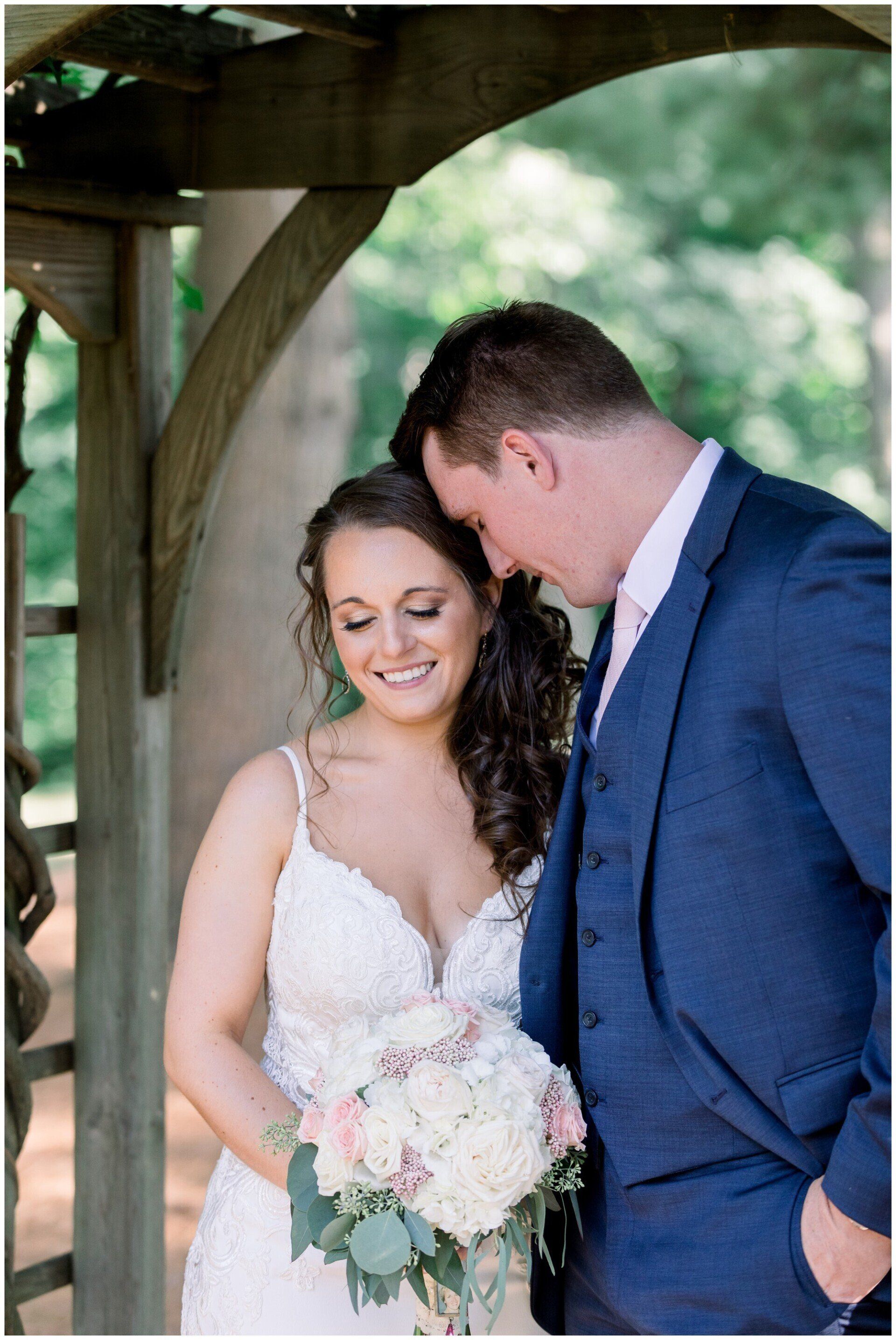 Bride and groom standing under a wooden pavilion, smiling and holding a bouquet at an outdoor wedding