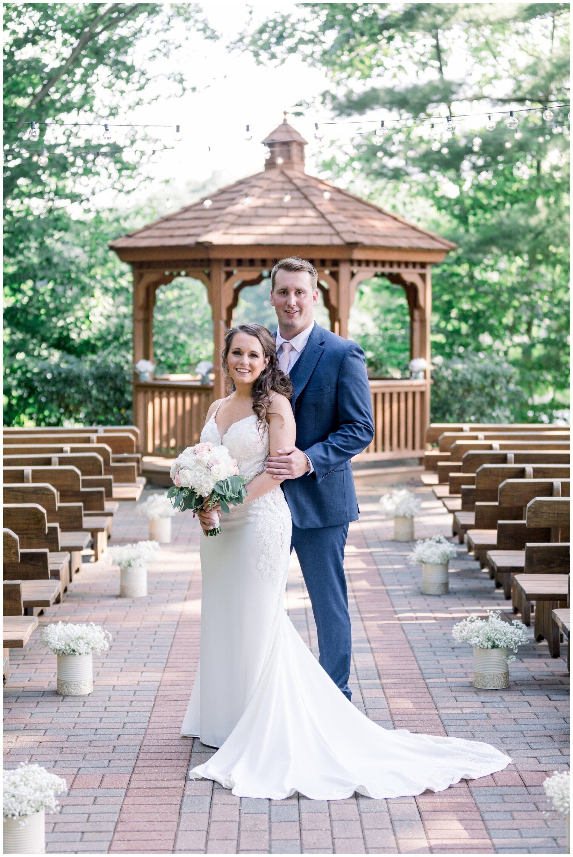 Bride and groom standing in an outdoor gazebo aisle, surrounded by empty chairs and greenery.