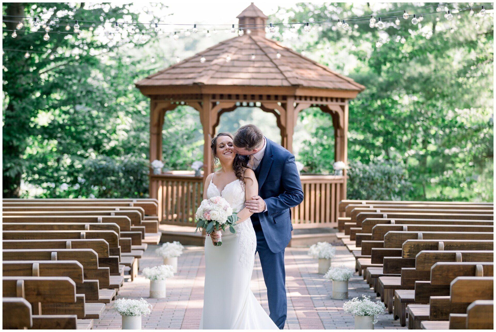 Bride and groom embracing in a gazebo aisle, surrounded by rows of wooden benches outdoors.