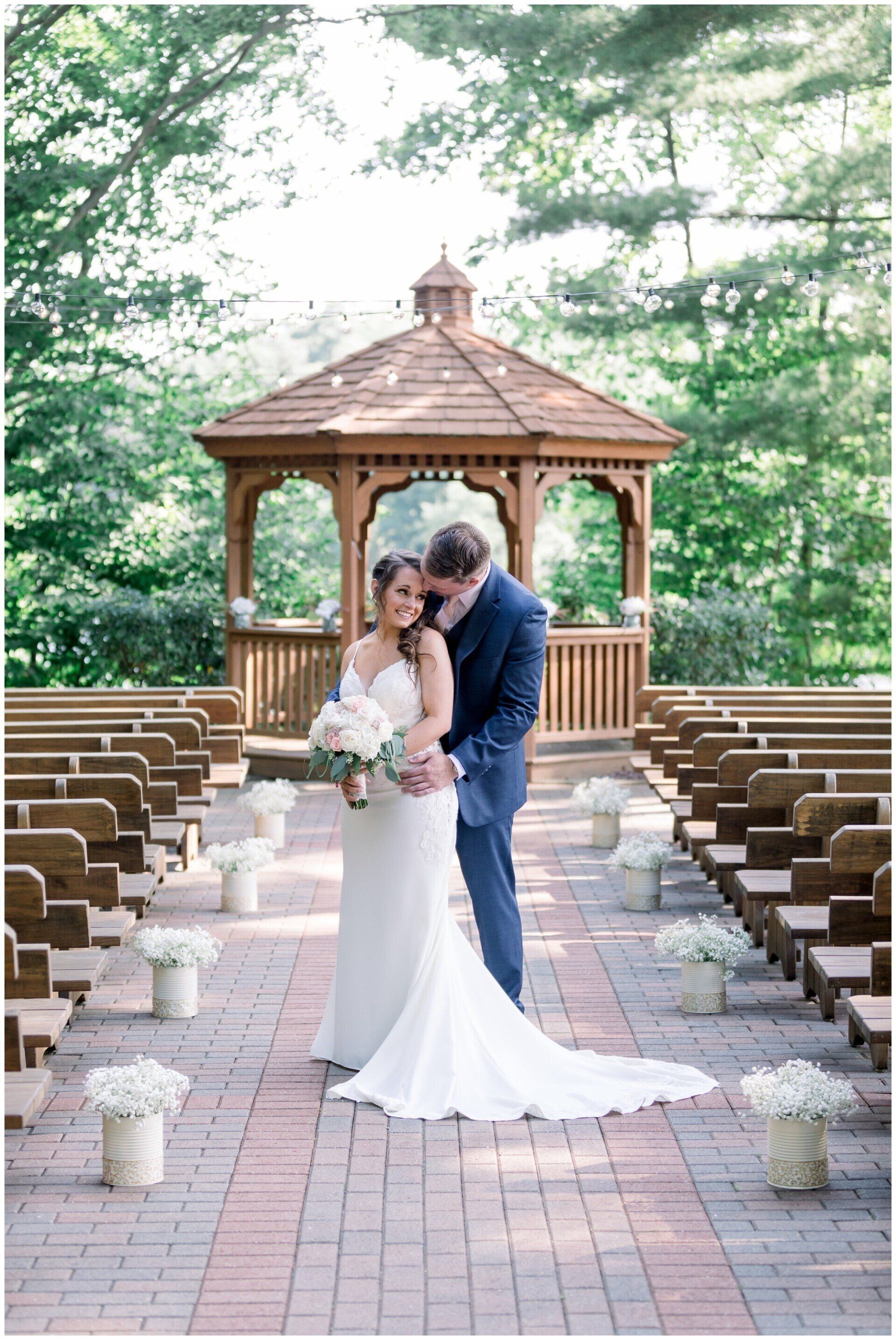 Bride and groom embracing in a garden aisle before a gazebo, with rows of benches and white floral accents