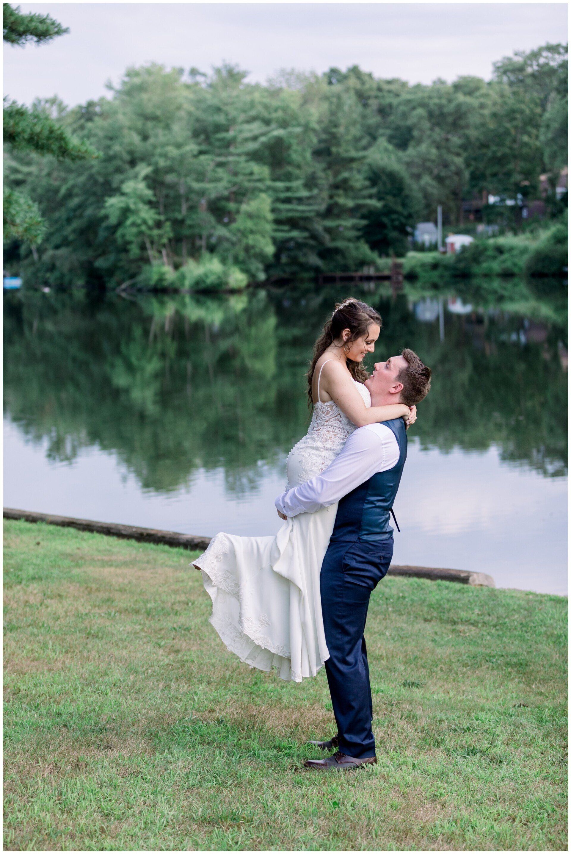 Couple embracing by a calm lakeside, with green trees reflected in the water.