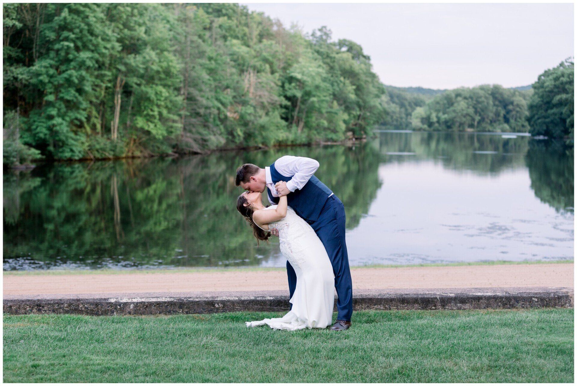 Bride and groom kissing by a calm lake, with lush green trees in the background.