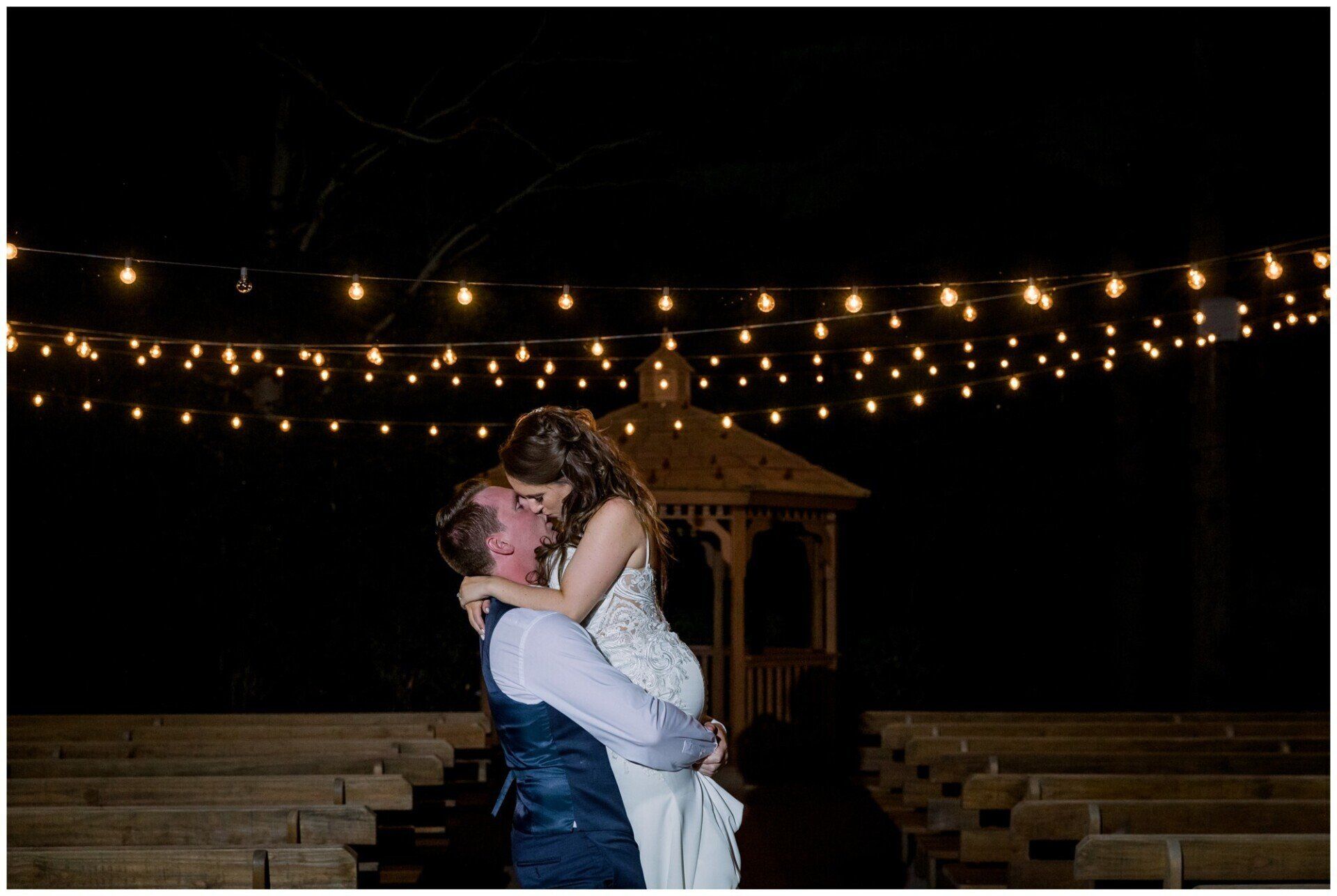 Couple embracing in a dimly lit outdoor venue with string lights and a gazebo behind them
