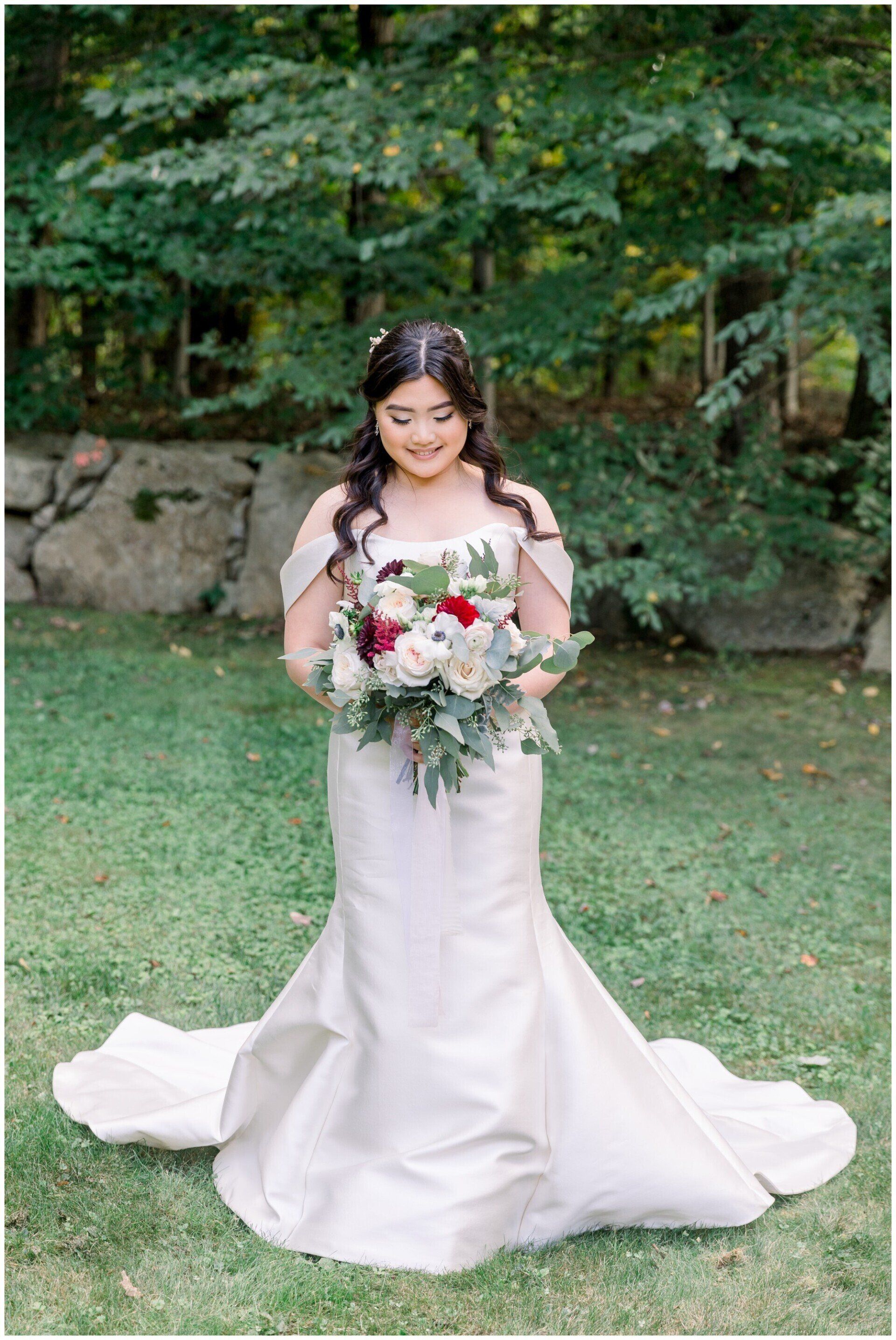 Bride in a white gown holding a bouquet, standing on grass in front of a wooded backdrop.