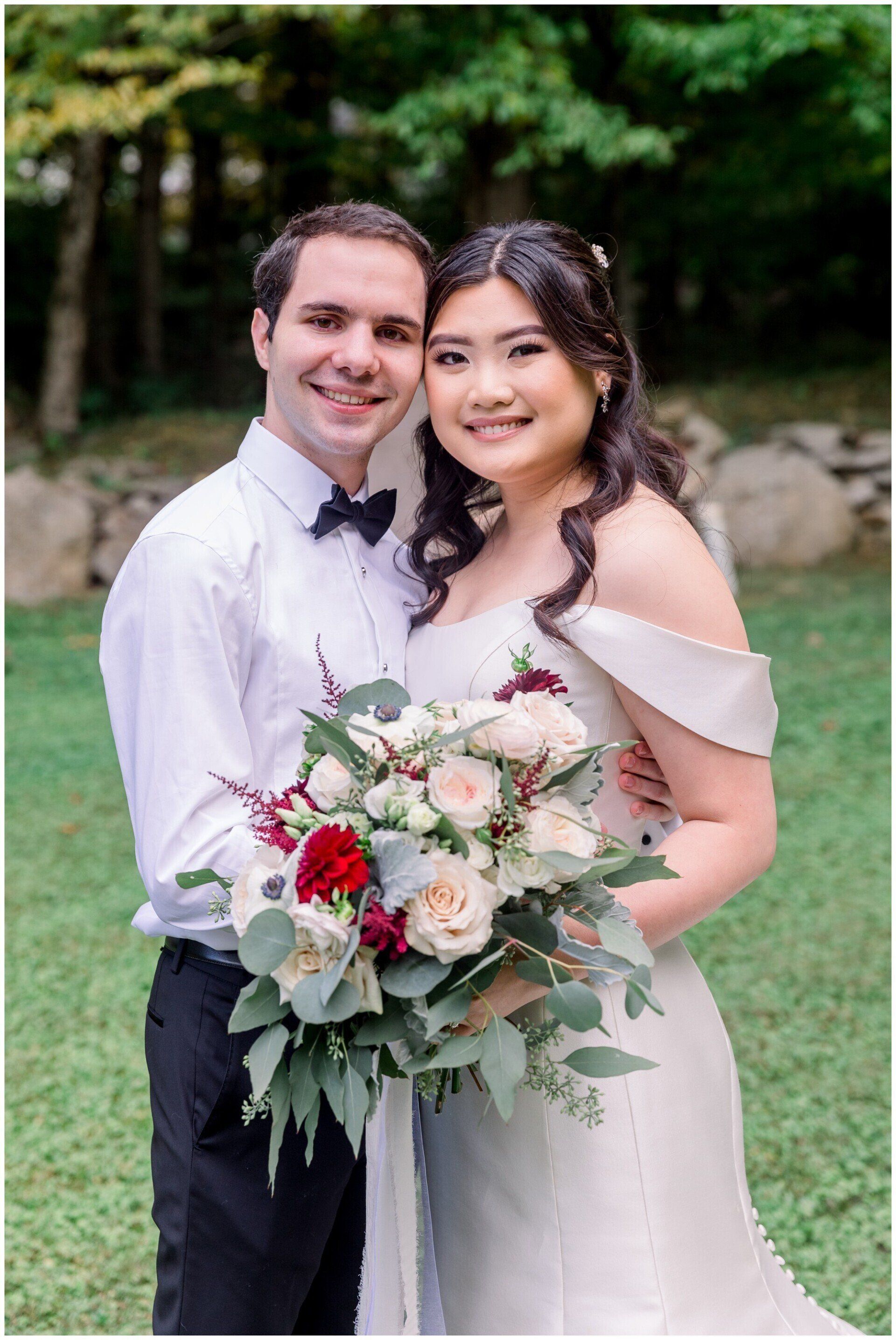 Newlyweds posing outdoors, holding a white and pink bouquet, smiling in wedding attire