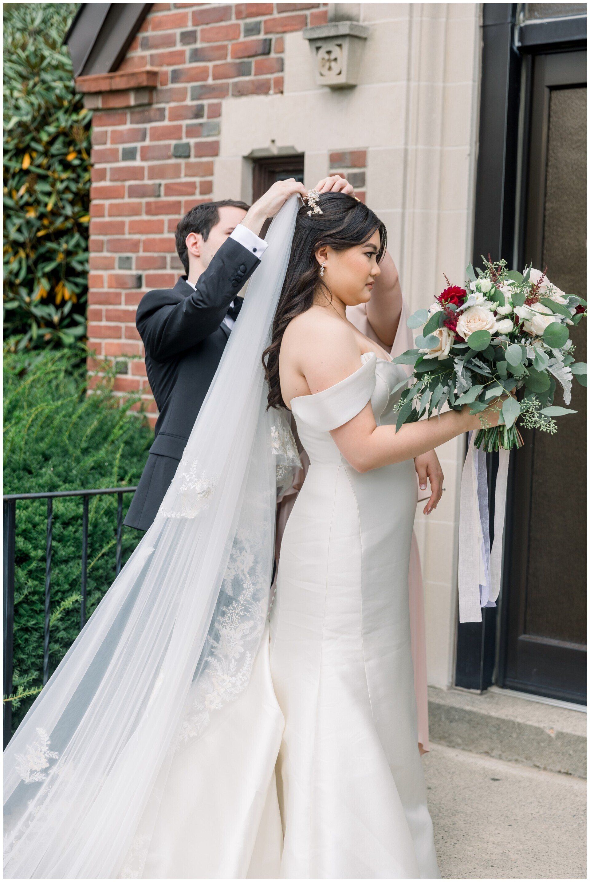 Bride with long veil and bouquet at a doorway while someone adjusts her veil outdoors