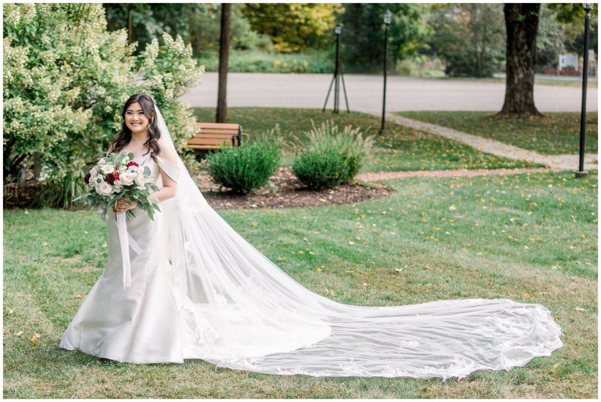 Bride in white gown holding a bouquet, standing on grass with a long veil trailing behind her in a garden
