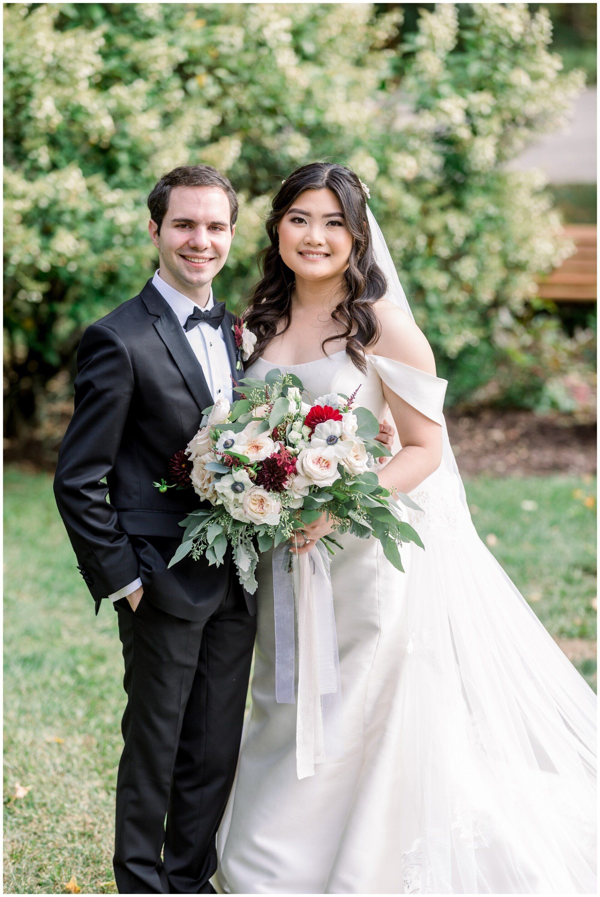 Bride and groom posing outdoors, holding a bouquet, in formal wedding attire