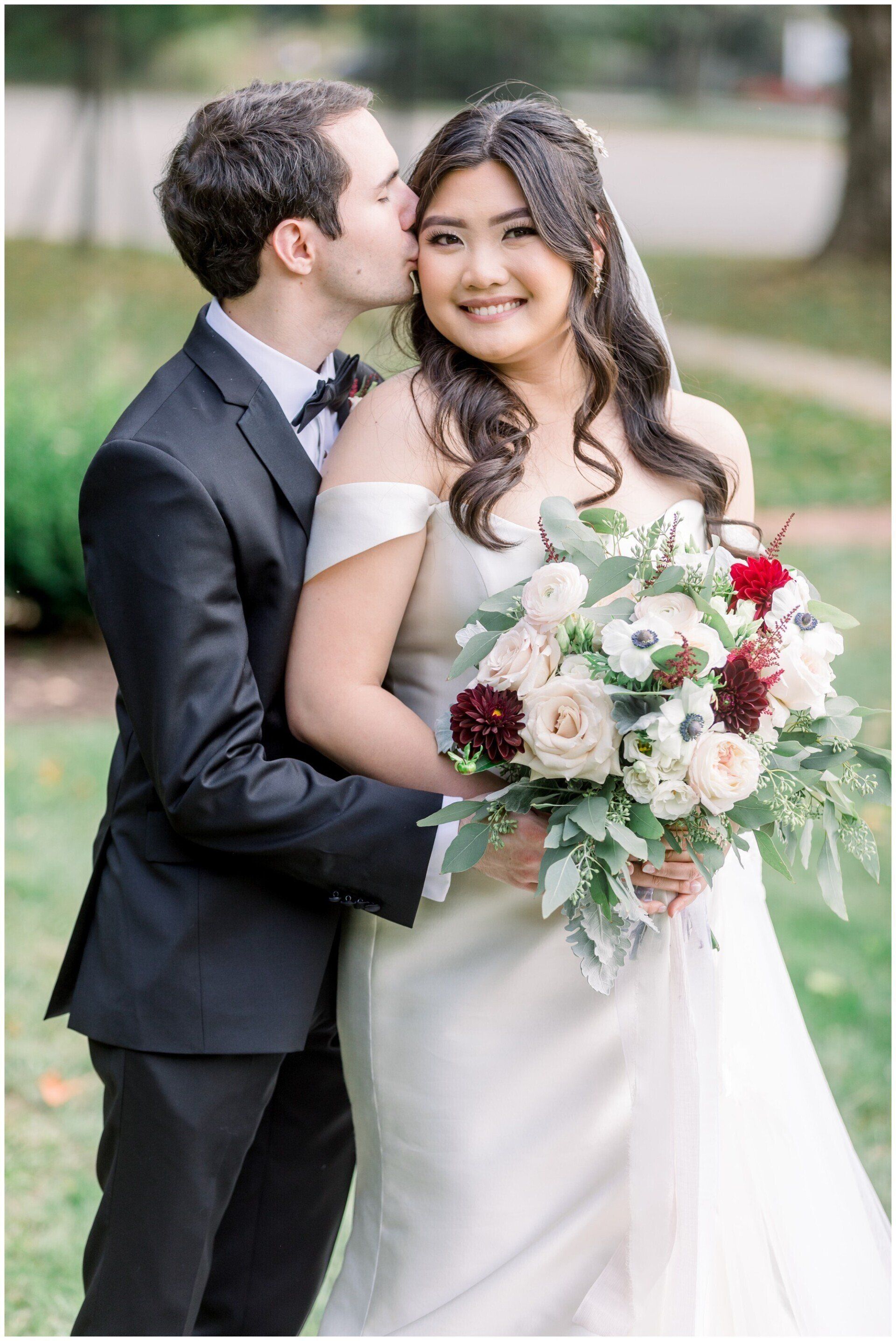 Bride and groom in formal attire, smiling outdoors as he kisses her cheek, holding a white and blush bouquet
