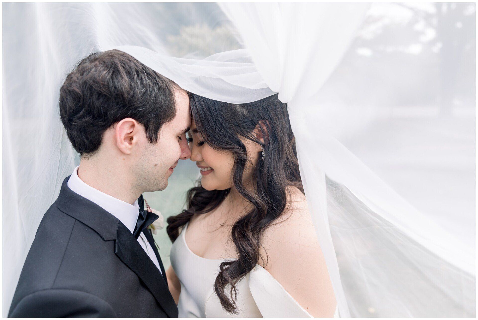 Bride and groom nuzzle under a white veil, smiling softly in a bright, airy setting