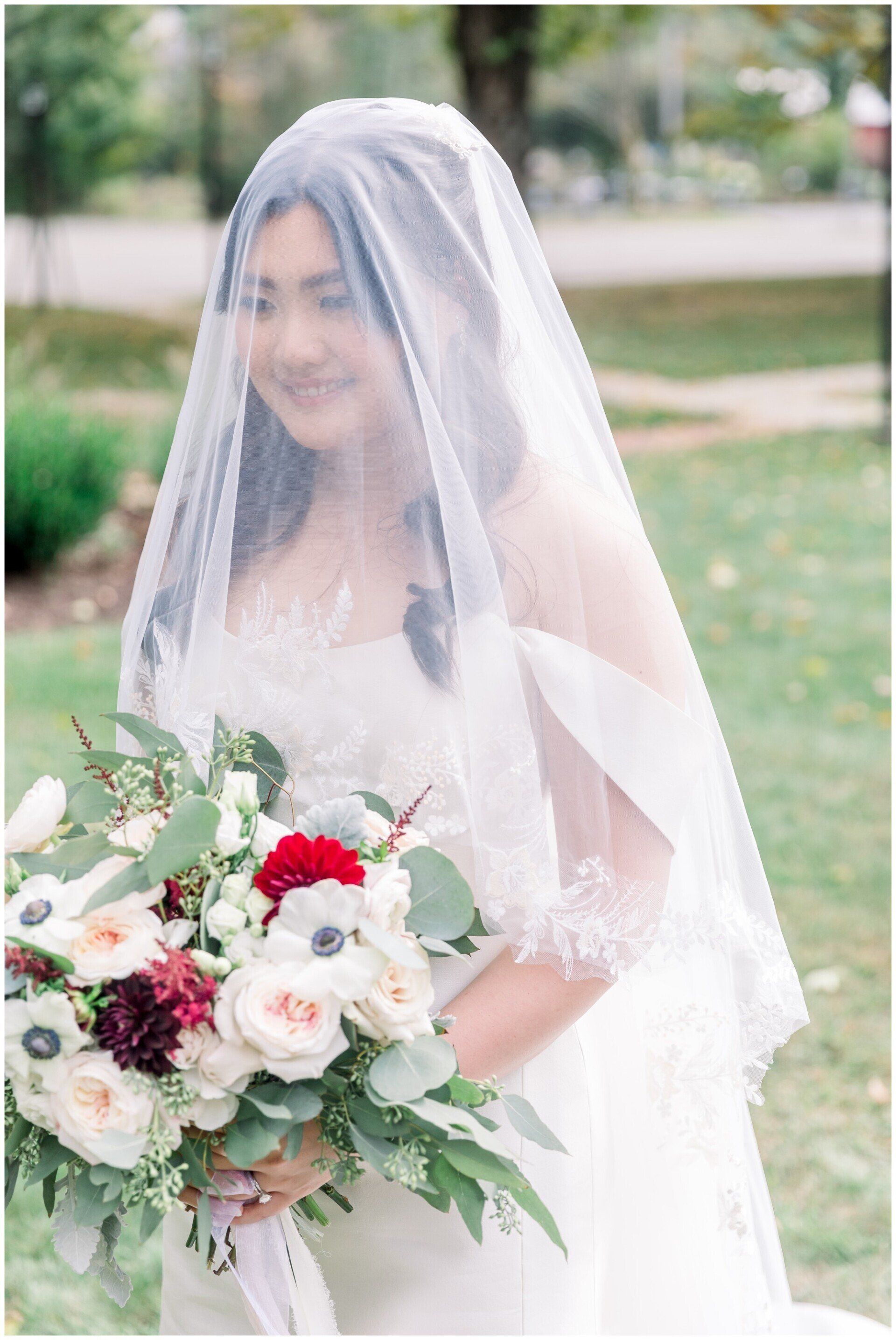 Bride in white veil holding a bouquet of white and red flowers outdoors