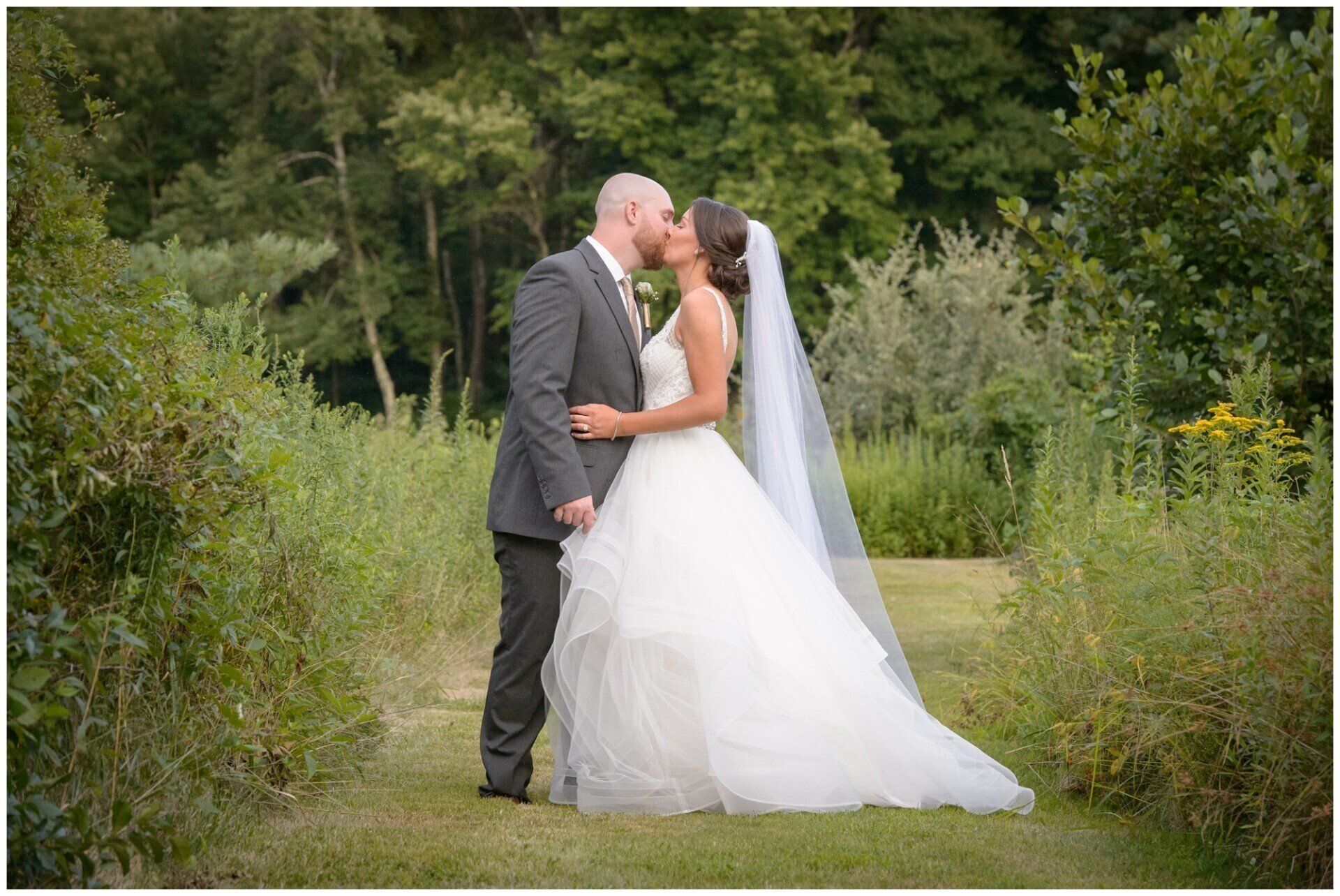 Bride and groom kissing on a grassy path in a lush green garden, wedding dress and veil visible