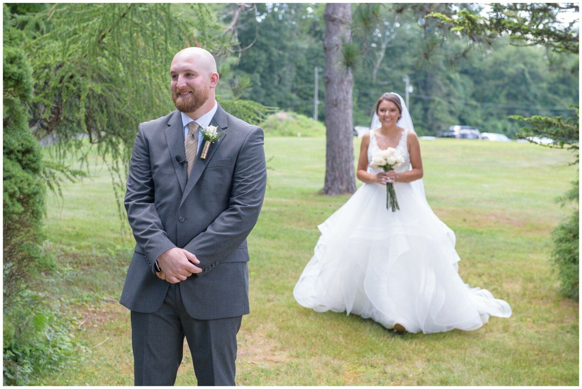 Groom in gray suit stands in foreground as bride in white gown walks behind him on a grassy lawn