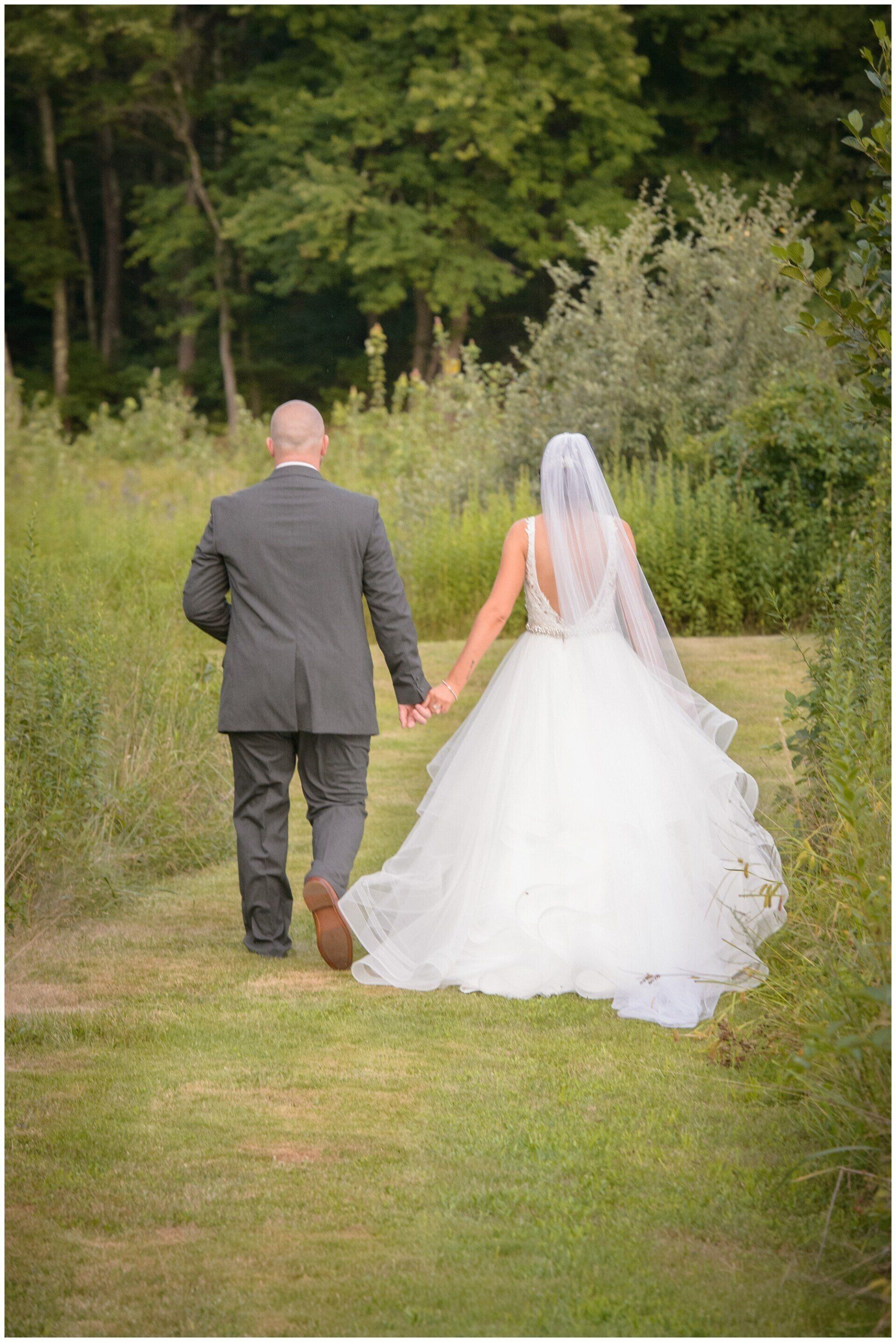 Bride and groom walking hand in hand down a grassy path in a garden, back view