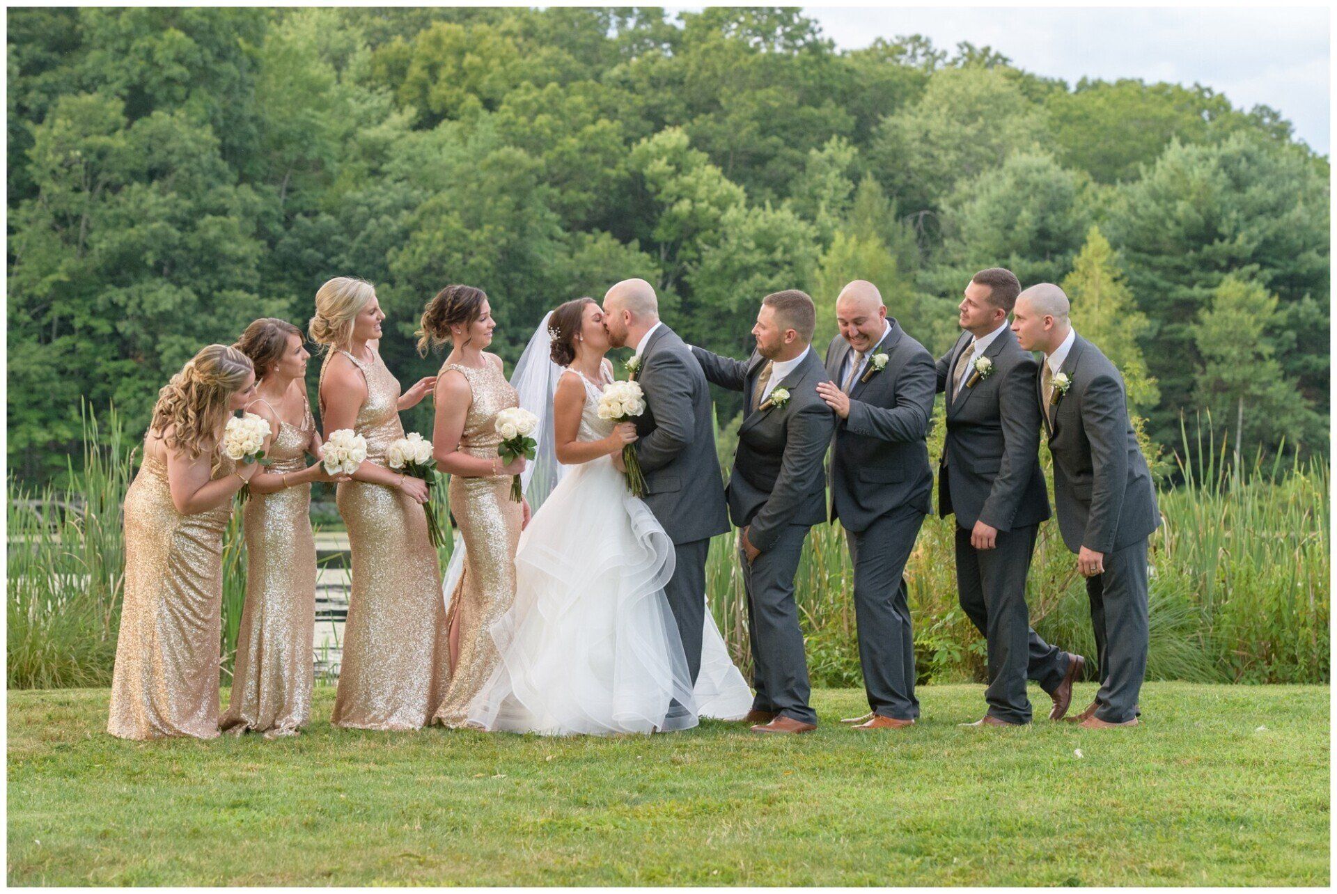 Wedding party in formal attire laughing outdoors beside a bride and groom in a grassy field
