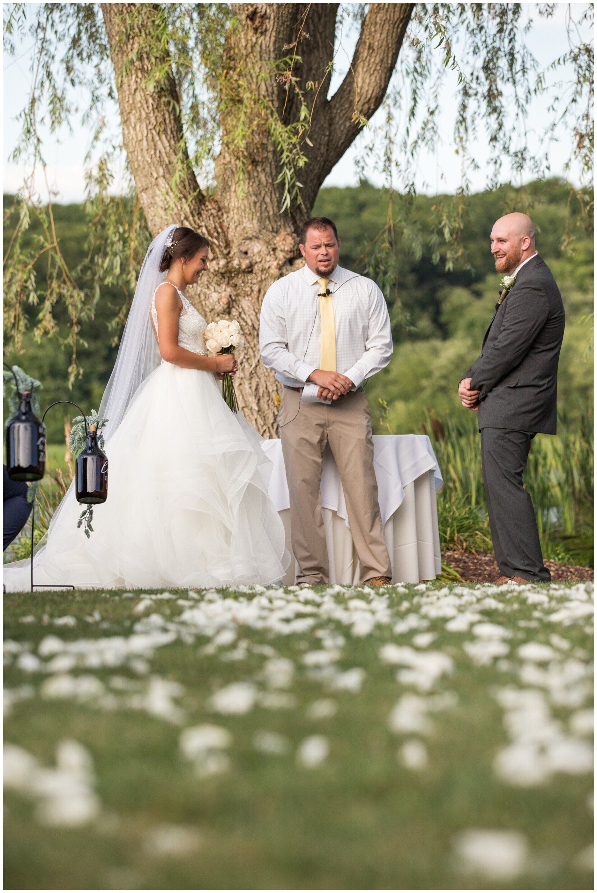 Bride and groom stand with an officiant outdoors beneath a tree during a wedding ceremony
