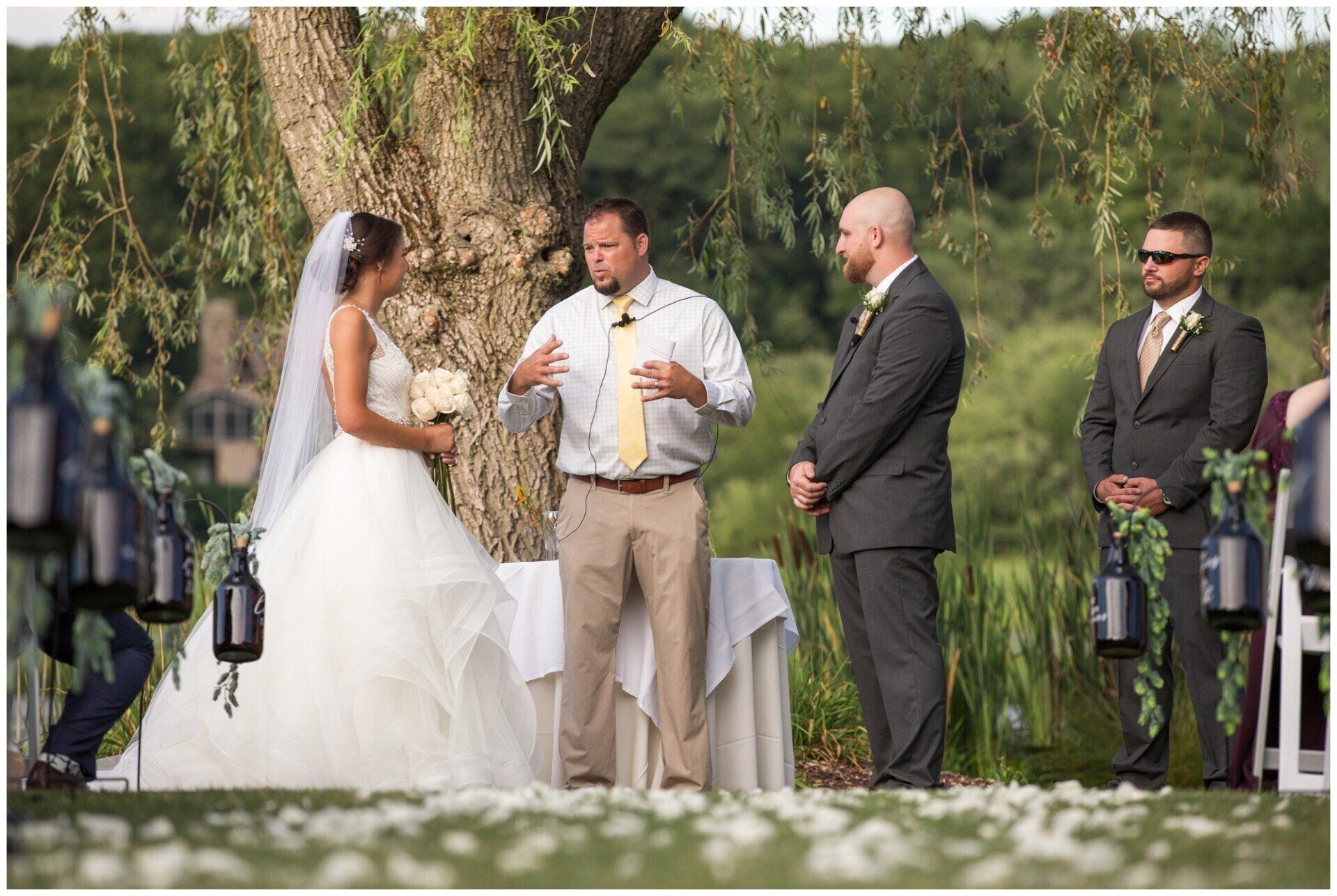 Outdoor wedding ceremony with bride and officiant under a tree, groom and groomsmen standing nearby