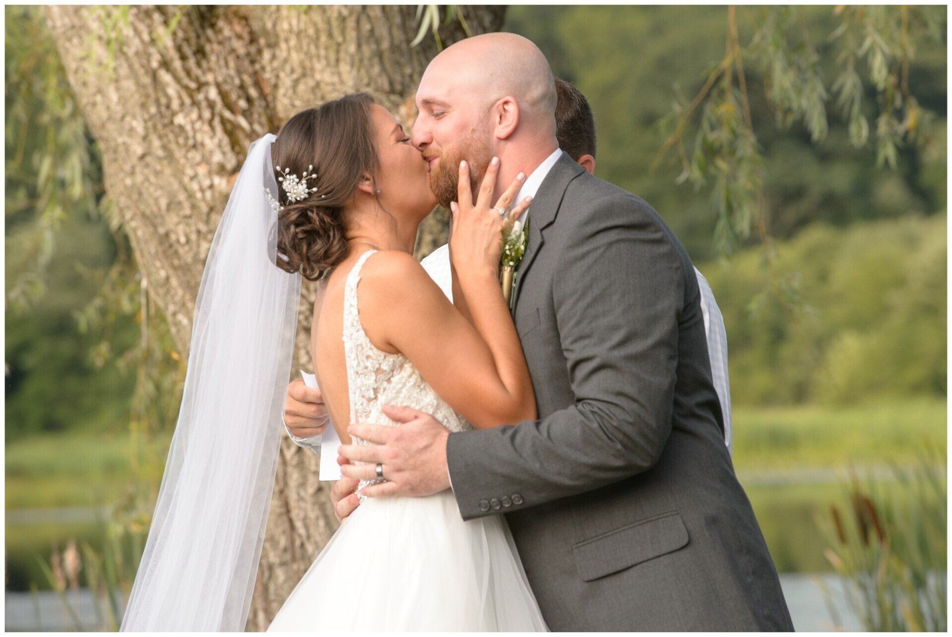 Bride and groom kissing beside a tree, with wedding dress, veil, and gray suit outdoors.