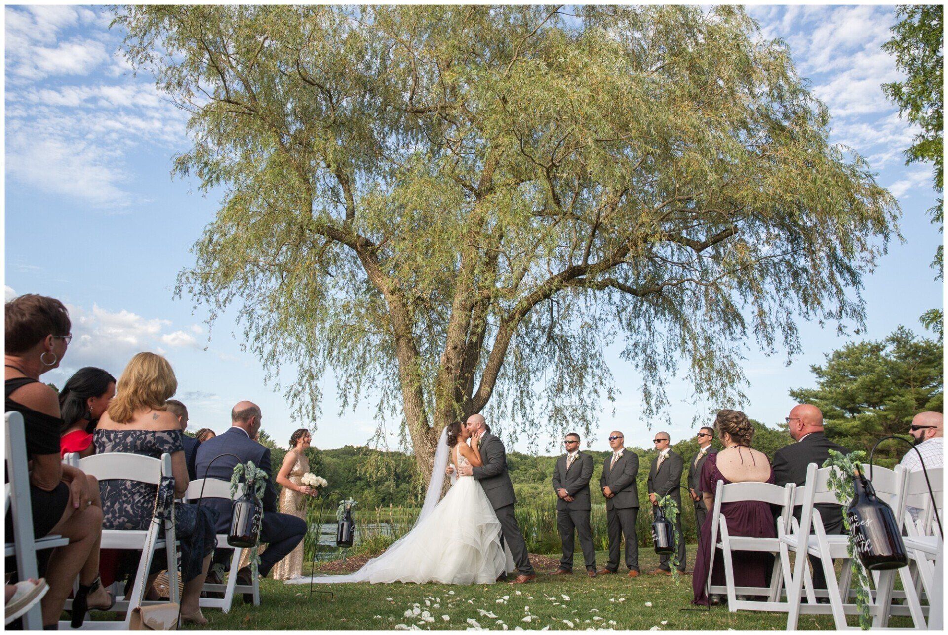 Wedding ceremony outdoors beneath a large tree, bride in white facing the groom and officiant, guests seated on both sides