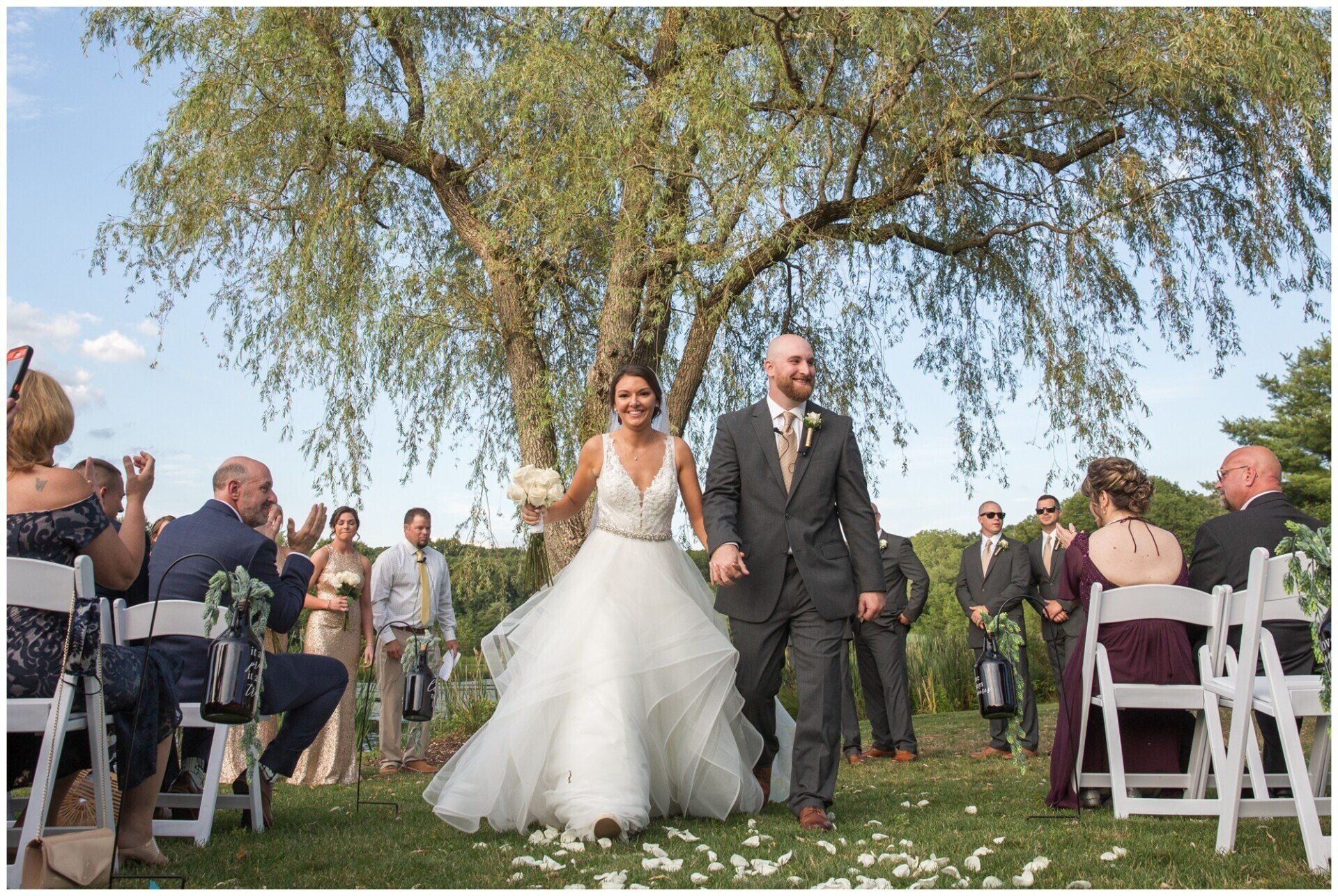 Bride and groom walk down an outdoor aisle under a tree at a wedding ceremony