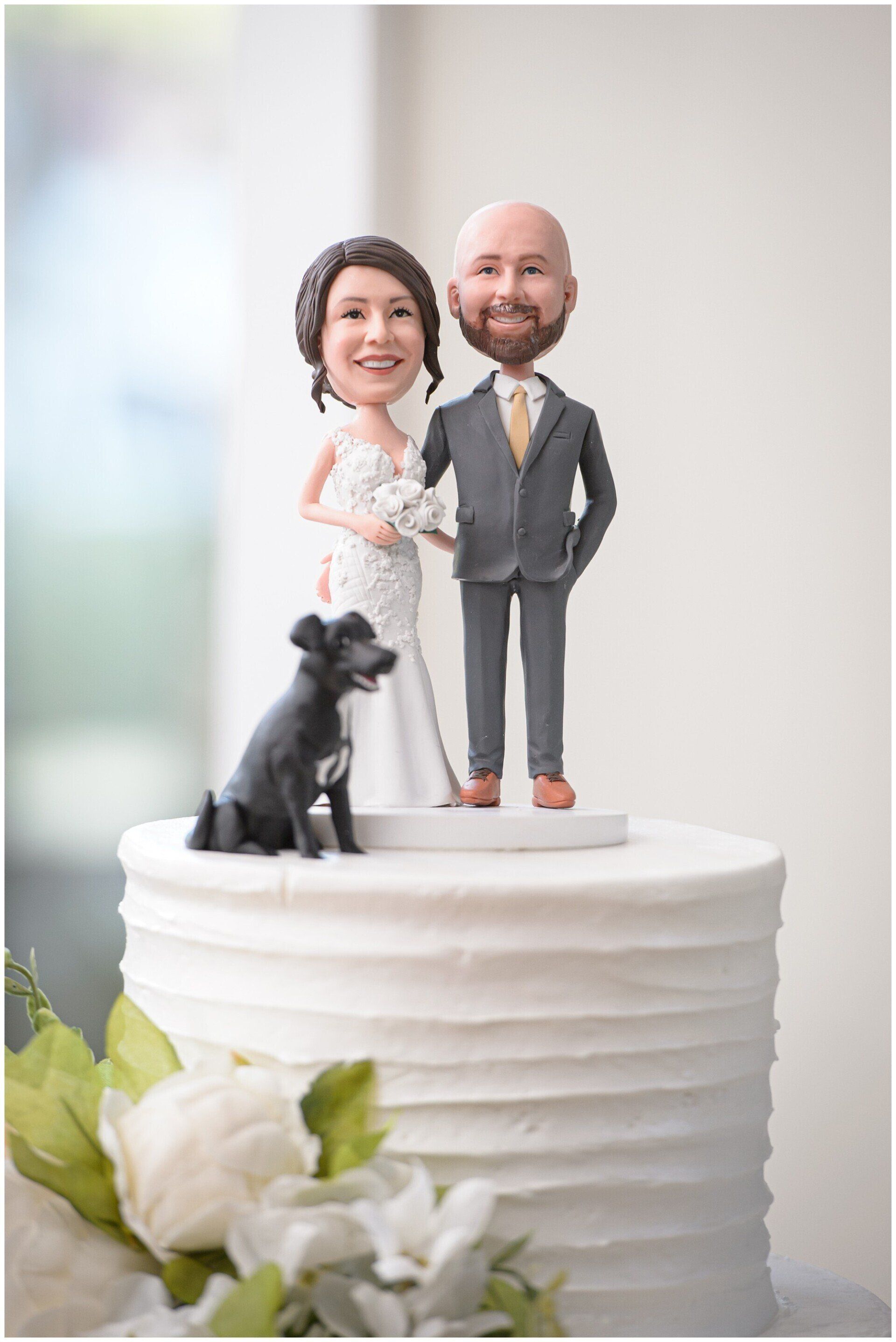 Wedding cake topper figurines with a dog on a white frosted cake, surrounded by white flowers.
