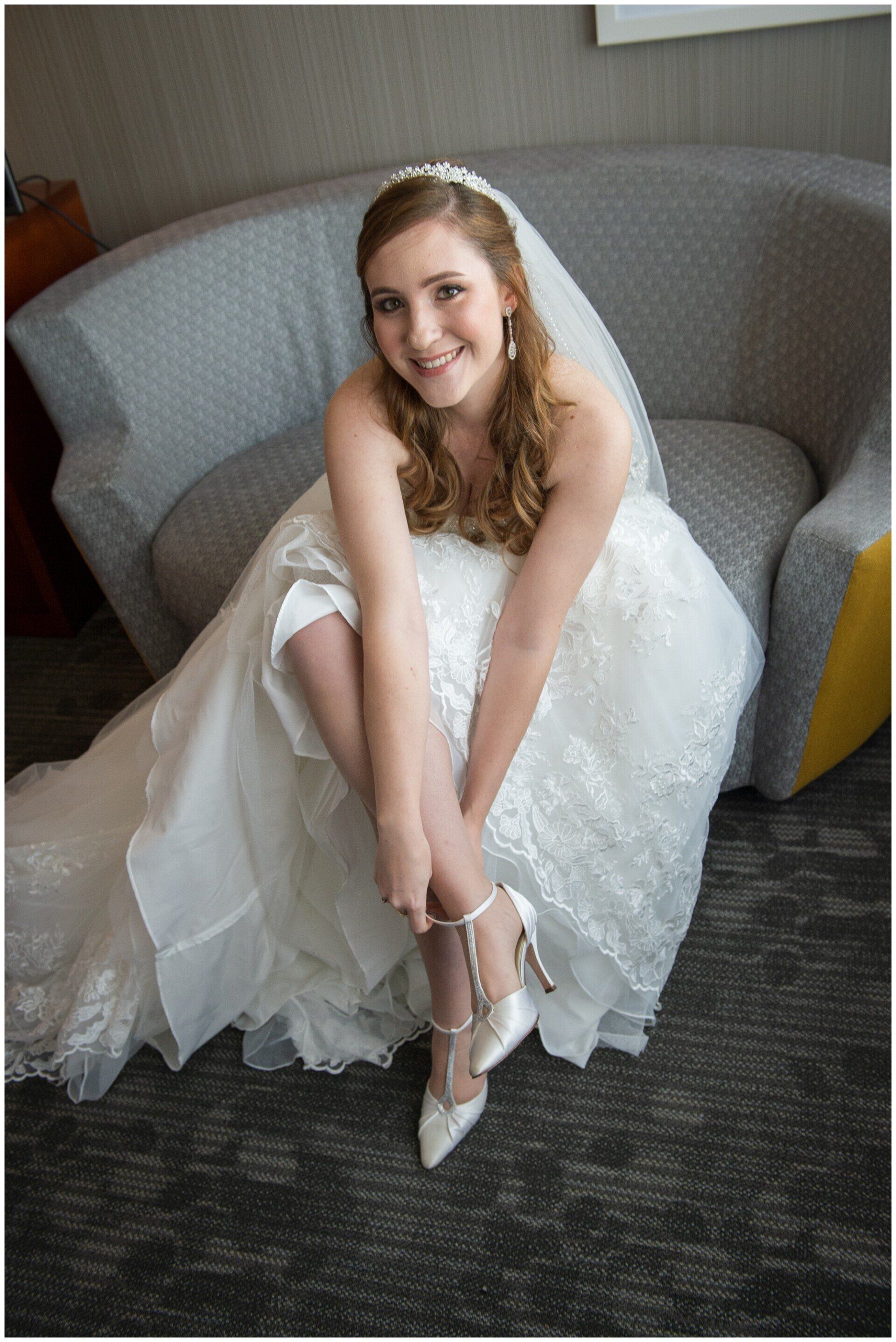 Woman in a white wedding dress sitting on a gray chair, smiling while fastening her silver heels.