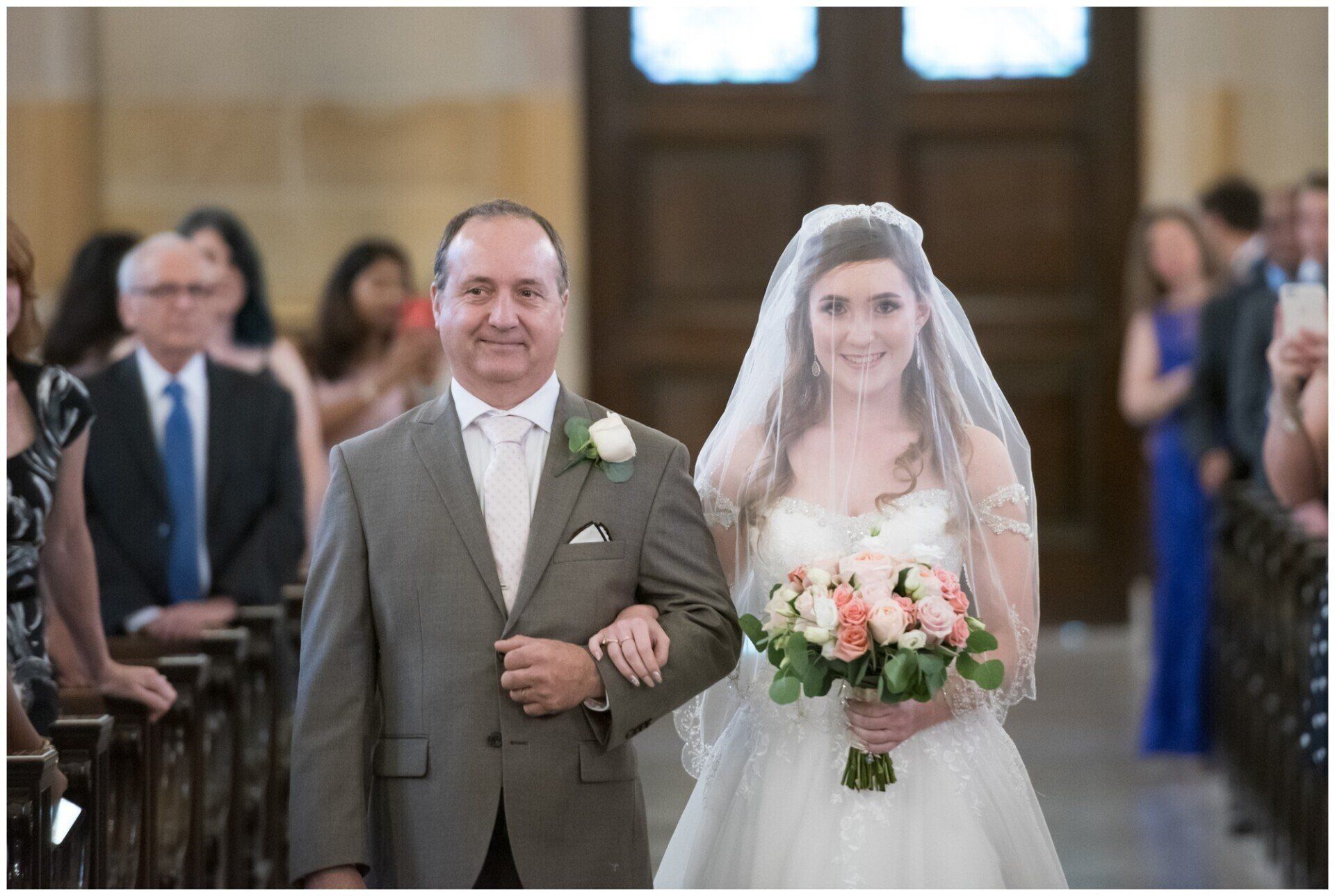 Bride walks down church aisle with a man in a suit, holding a bouquet and wearing a veil.