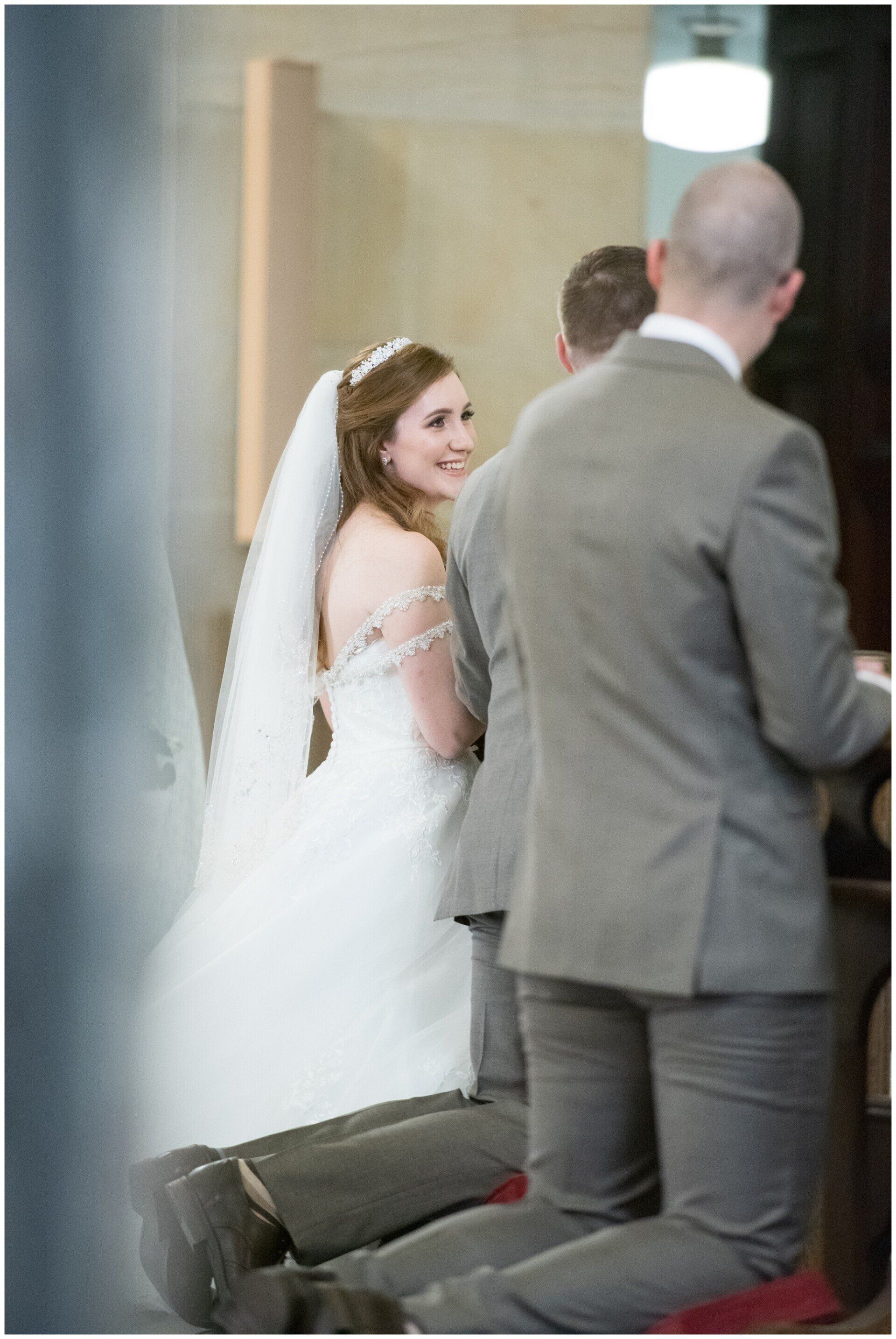 Bride in a white veil glancing back during a wedding ceremony, with a man in a gray suit in the foreground