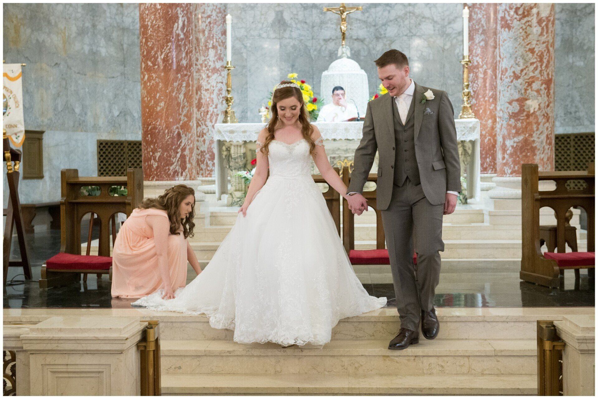 Bride and groom walking down church steps, with a woman kneeling beside them in a wedding ceremony
