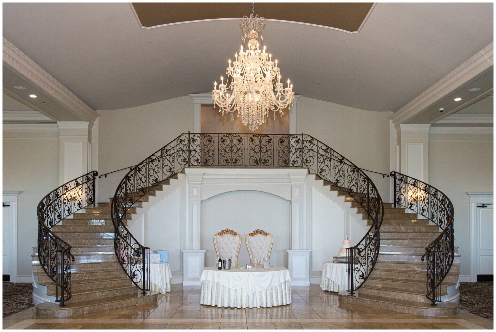 Grand ballroom with a crystal chandelier and twin curved staircases framing a central table.