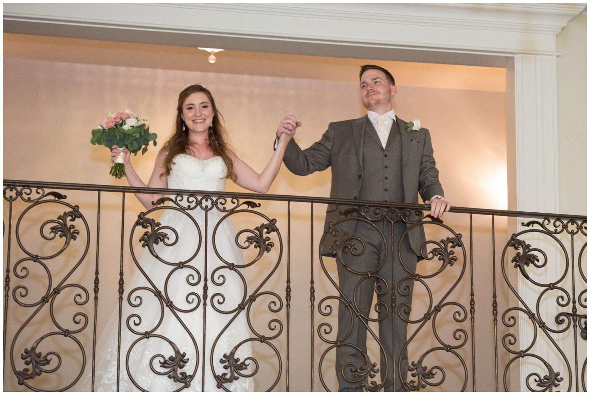 Couple standing on a balcony, holding hands, with a bouquet of flowers nearby.