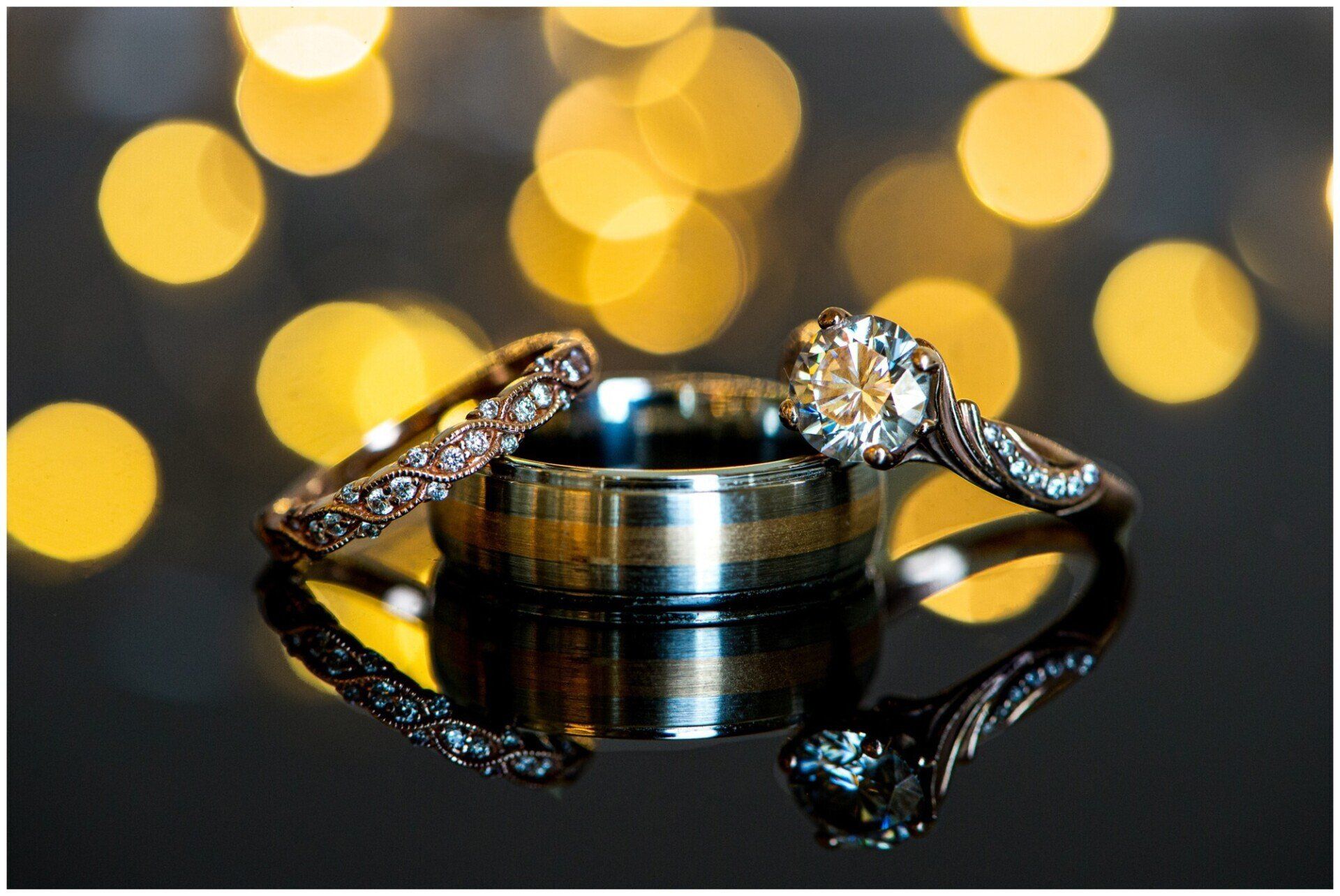 Two wedding rings on a reflective black surface with blurred warm lights in the background